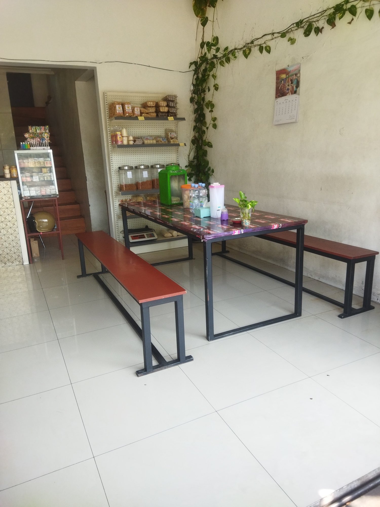 The image depicts the interior of a food stall or small restaurant located in Giripurwo, Indonesia. The room is brightly lit with natural light and features tiled flooring. A long, rectangular table with a colorful, patterned surface is centrally placed, supported by black metal legs. Two long, dark-colored benches with red seats are positioned along the sides of the table.
To the left of the table, a shelf unit holds various food items in clear jars and some packaged snacks. A green appliance, possibly a cooler or dispenser, is also visible on the shelves. In the background, a staircase leads to an upper level, and a display refrigerator contains more products. A helmet rests on a stand near the staircase.
On the wall behind the table, a calendar is hanging, and a vine plant trails along the wall and ceiling, adding a touch of greenery to the space. Several bottles of water and a plant in a glass vase are on the table. The overall impression is that of a simple, functional establishment ready to serve customers.