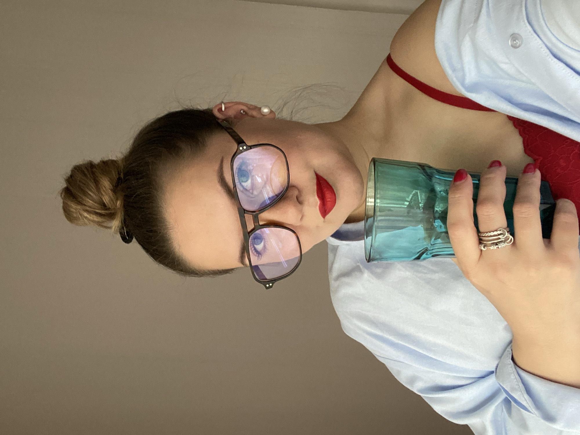 A woman with her hair in a bun, wearing glasses, red lipstick, a red bra, and a blue button-down shirt, is holding a blue glass. The location is not discernible as Izhevsk, Russia, or any other specific location.