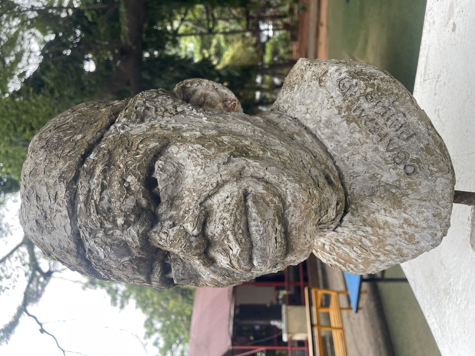 A low-angle, close-up shot depicts a weathered, light-colored stone bust of a human head. The head appears to be wearing a hat or head covering. The bust shows a significant degree of texture, likely due to age and environmental exposure. The sculpture is placed outdoors, with trees visible in the blurred background. A building with red and yellow framing can be seen in the lower portion of the image, suggesting the location is likely within a park or garden setting in an urban environment. The bust itself exhibits possible inscription; a close examination of the stone reveals the words "OCHETEU 83 Vasile."
