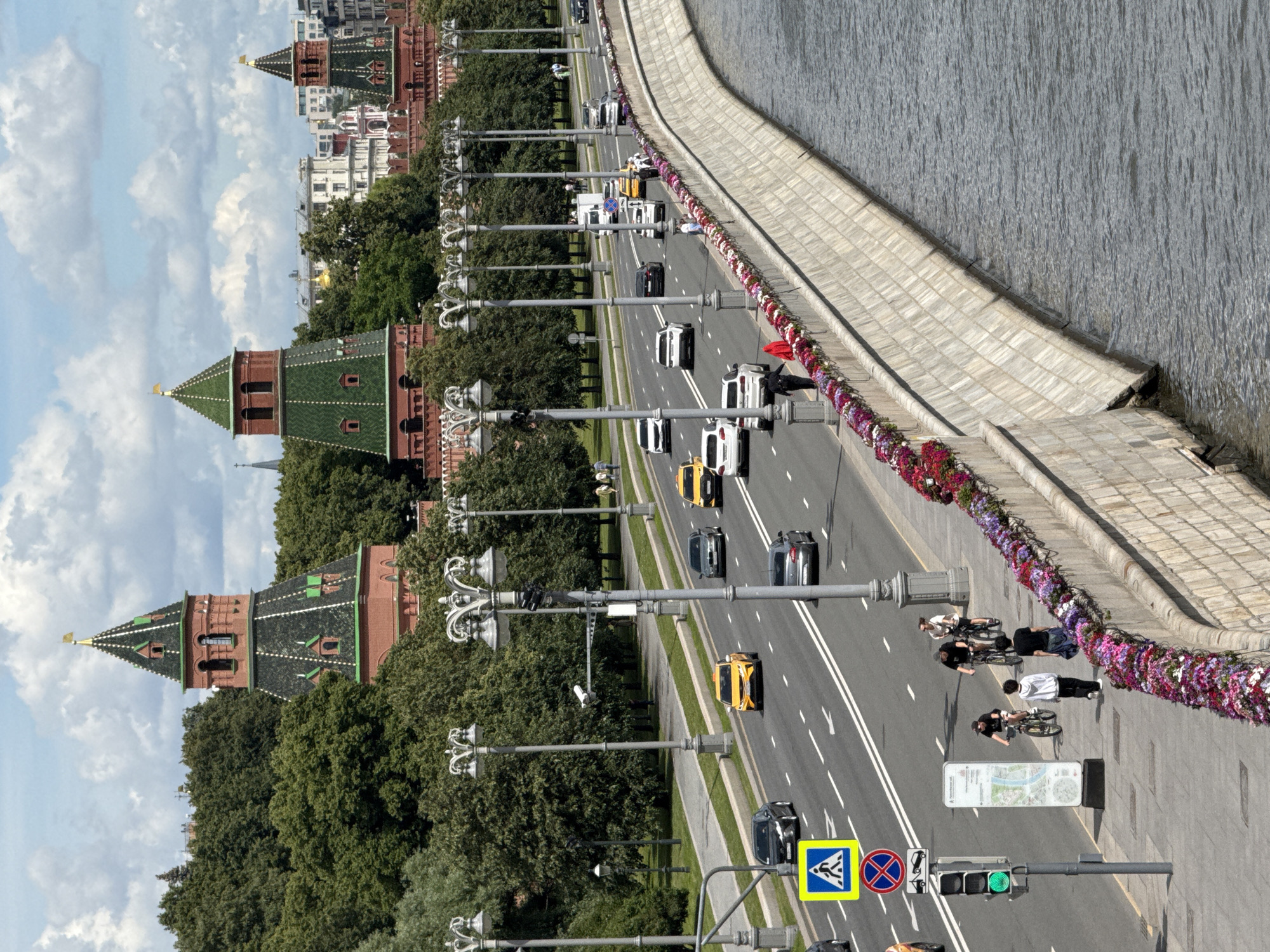 The image depicts a wide, curved river embankment lined with a stone wall and a walkway. Along the embankment, a row of flowering plants in various shades of pink and purple adds color. A multi-lane road runs parallel to the embankment, with several cars and taxis driving on it. Traffic lights and street lamps are visible along the road. On the walkway, a few pedestrians and cyclists are present. In the background, lush green trees partially obscure imposing red brick towers with green roofs, characteristic of the Moscow Kremlin. The sky above is partly cloudy. The scene appears to be a bustling urban environment in Moscow, Russia, likely a popular tourist area.