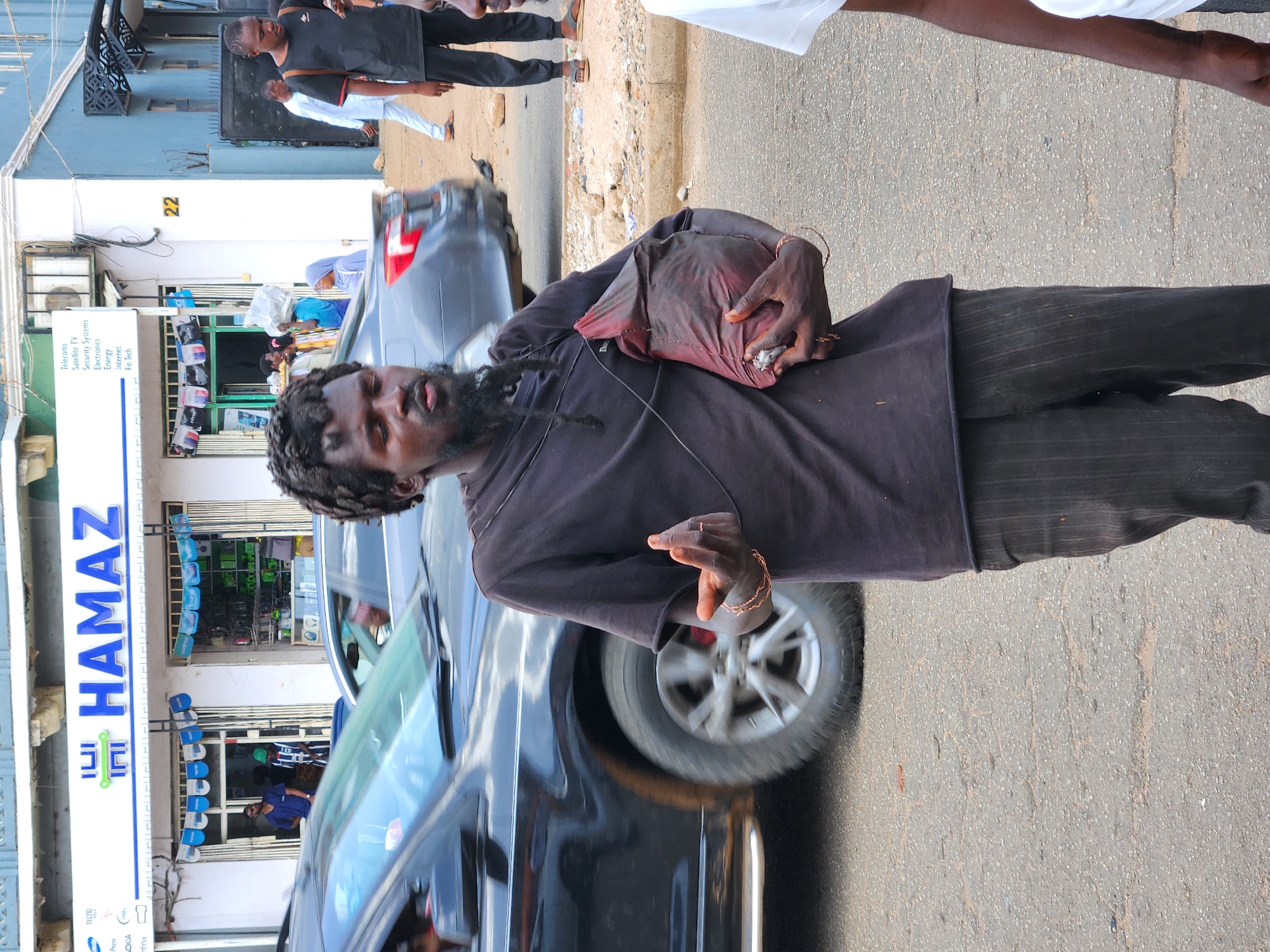 The image shows a man with dreadlocks standing on a city street in front of a business with a sign that reads "HAMAZ," offering services such as telecoms, satellite TV, security systems, electronics, energy, internet, and Fin Tech. He is holding a reddish-brown bag over his shoulder, has a bracelet on his right wrist, and is gesturing with his right hand. A black car is partially visible in the foreground. Other people can be seen walking in the background near the business. The building next to the business has the number "22" displayed on it. The street appears to be paved and in average condition.