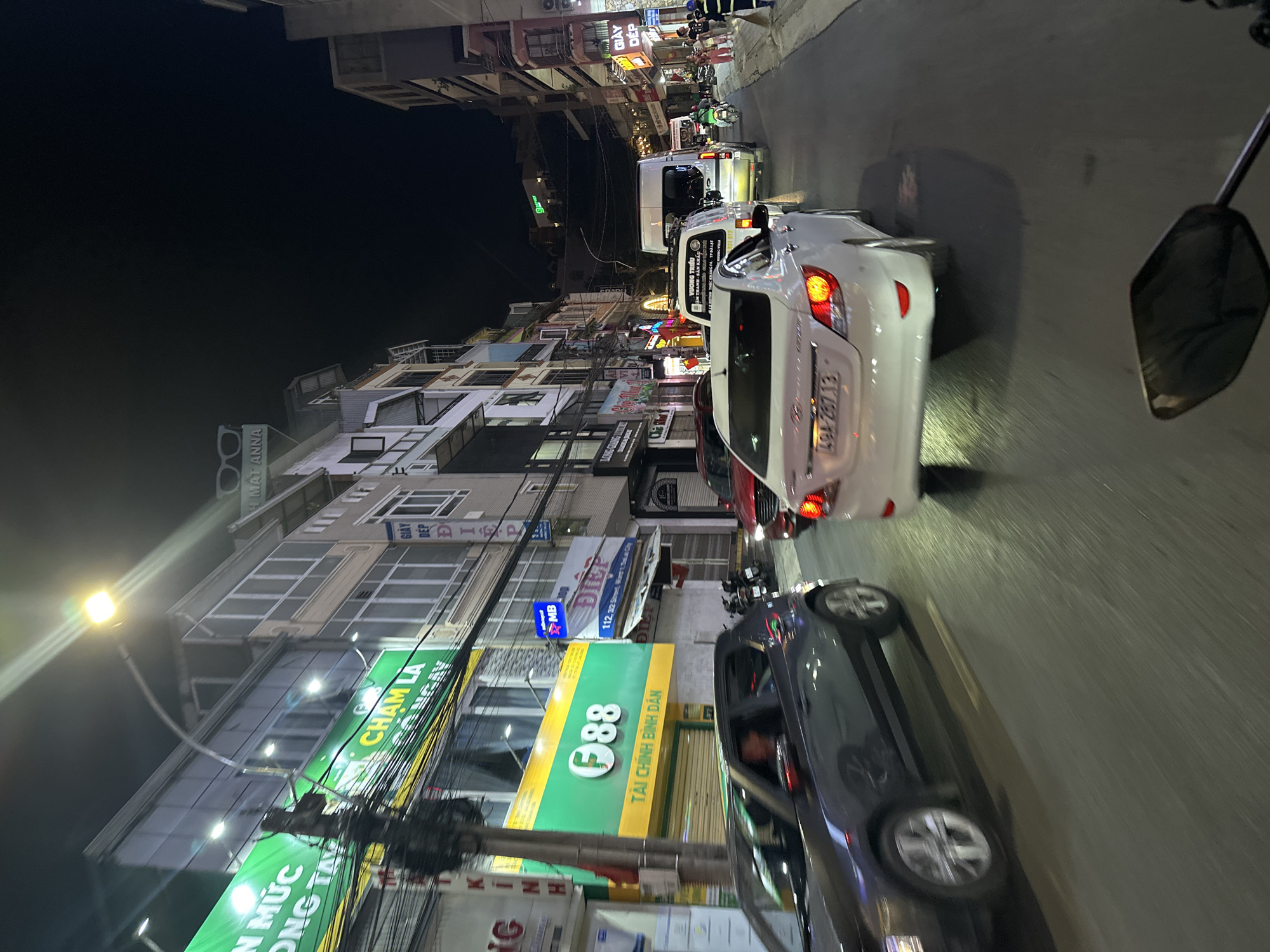 This media file captures a nighttime scene on a street in Da Lat, Vietnam. The perspective is from a moving vehicle, likely a motorcycle, looking forward.
The main subjects are cars and buildings lining the street. Several cars are visible, including a white sedan with its taillights illuminated and a dark-colored sedan in the foreground. The buildings are multi-story structures with businesses on the ground floor, indicated by illuminated signs. There are also some people visible, mostly as passengers or drivers within the vehicles, and a few pedestrians can be seen on the sidewalk.
The activity is the movement of traffic at night. The street appears to be congested, suggesting a busy urban environment. The time of day is clearly night, as evidenced by the darkness and the illuminated signs and vehicle lights. The weather is not explicitly indicated but appears to be clear enough for driving.
Notable details include the vibrant neon signs of businesses, some of which display text in Vietnamese and English. Prominent signs include "CHAM LA," "88 TÀI CHÍNH BÌNH DÂN" (indicating a financial service), "MB," and "GIÀY DÉP" (shoes). The license plate of the white sedan reads "494-267.13." The presence of a street lamp in the upper left quadrant also contributes to the nighttime ambiance. The overall mood is one of urban activity and the bustle of a city at night.
The location is confirmed to be Da Lat, Vietnam, with specific street references visible on some signs.