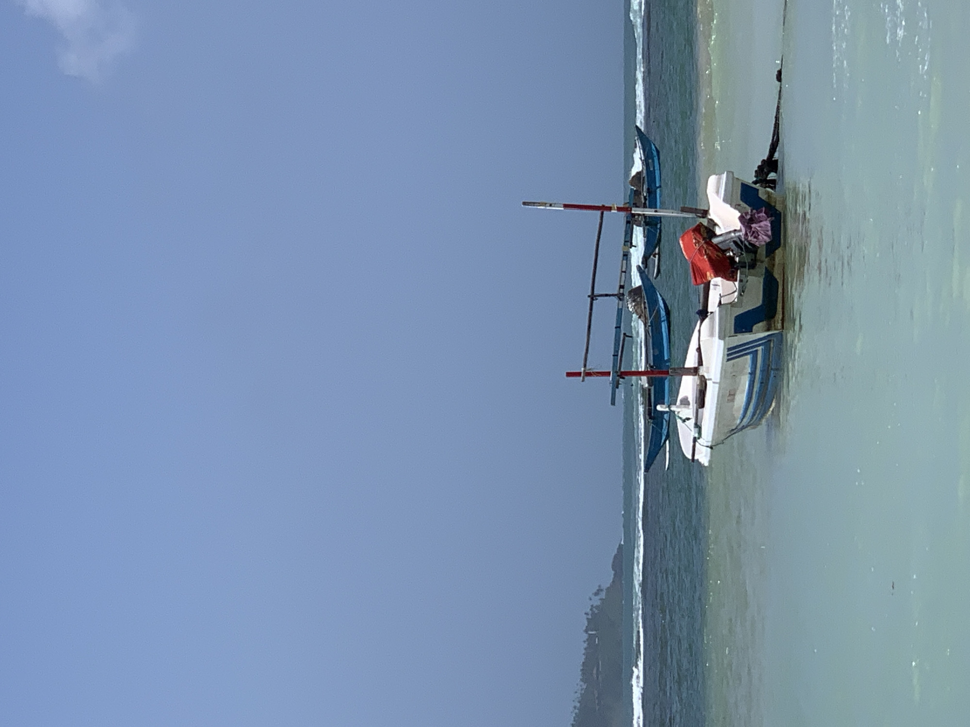 The image depicts a serene coastal scene in Weligama, Sri Lanka. In the foreground, a small, white and blue boat is anchored in calm, shallow, turquoise water. It has a red object secured to its stern, likely a fuel can, and a purple cloth draped over its engine. A thick rope extends from the boat to the right, disappearing into the water.
In the middle ground, two larger, traditional blue fishing boats are moored further out in the sea. They appear to be equipped with poles for their outriggers and have nets or other fishing gear covered on deck. Gentle waves are visible breaking in the distance.
The background shows a lush, green headland dotted with buildings and palm trees, characteristic of the Sri Lankan coastline. The sky is a pale, overcast blue, suggesting a slightly hazy or overcast day. The overall impression is one of tranquility and a typical fishing village setting.