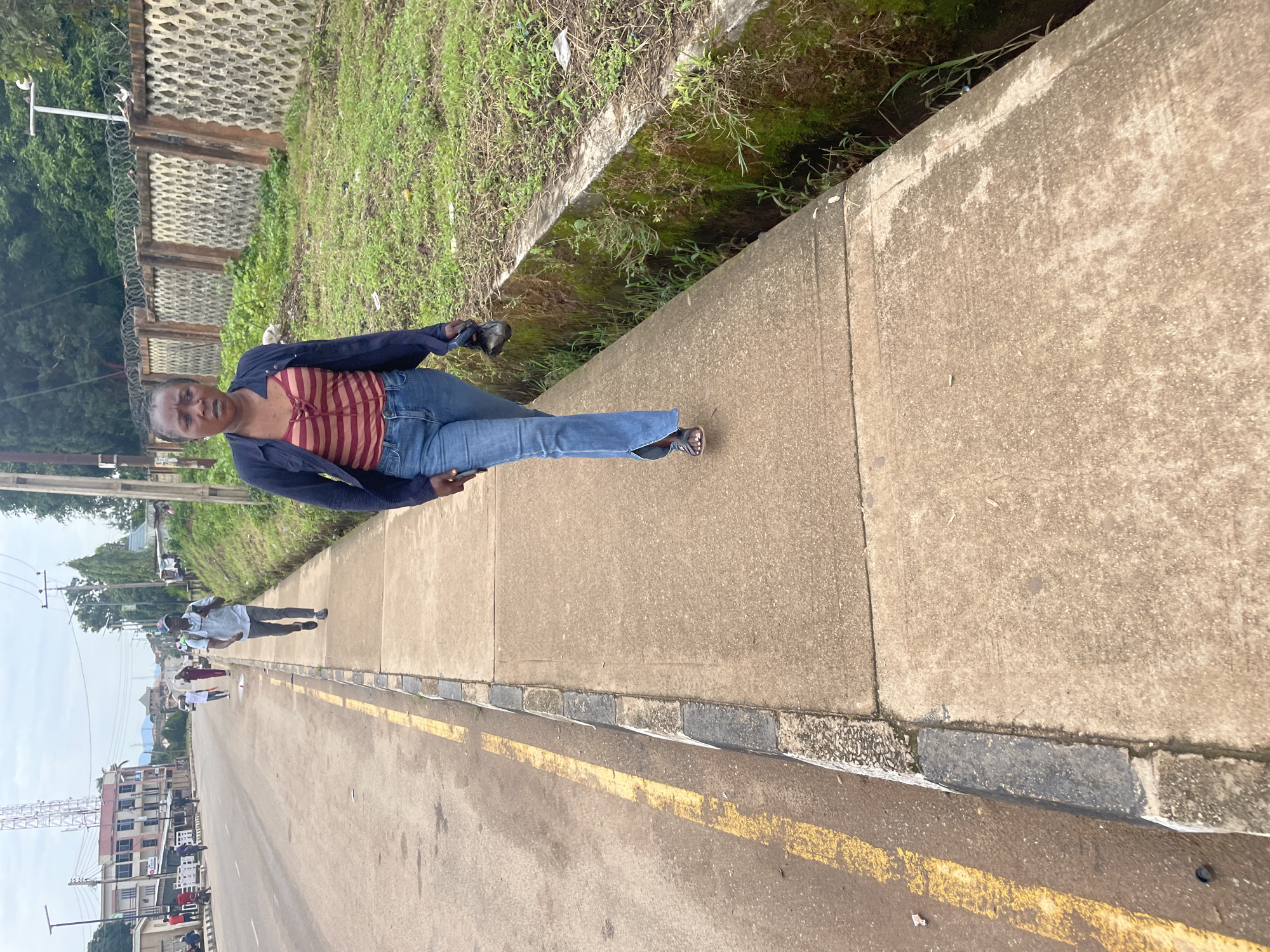 A Black woman walks on a concrete sidewalk next to a street in Jos, Nigeria. She is wearing a blue jacket over a red and white striped top, blue jeans, and black sandals. She is holding a black bag in her right hand. The sidewalk is bordered by a grassy area and a concrete drainage ditch. The street is paved and has a yellow line on the left side. In the background, there are buildings, trees, and a wall. There are also other people walking on the street and sidewalk in the distance. The sky is overcast.