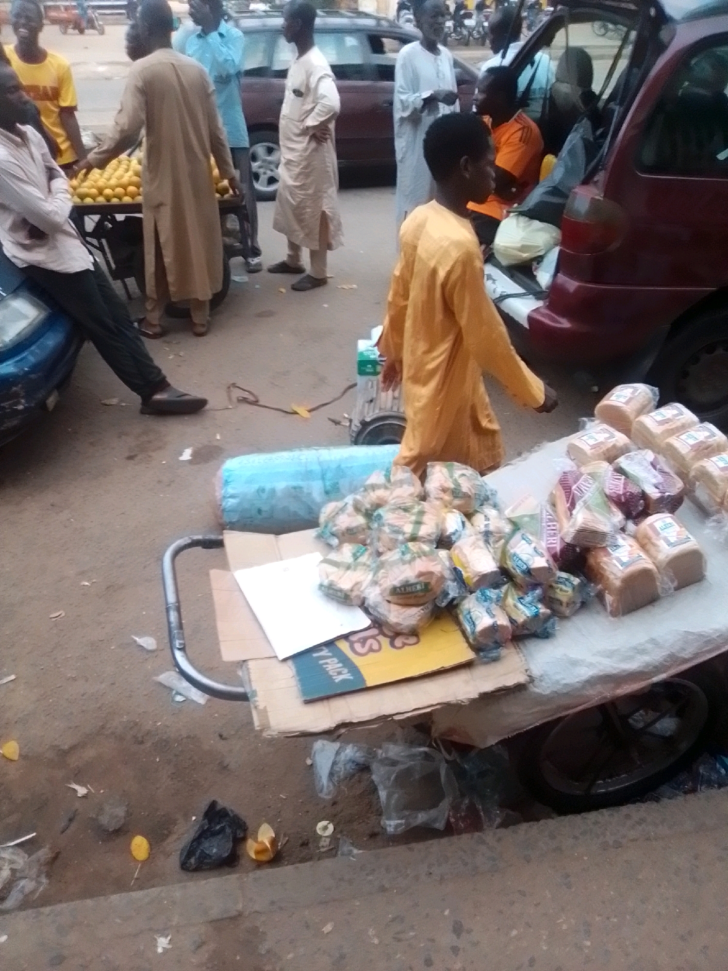 The image depicts an outdoor market scene in Arawa, Nigeria. In the foreground, a vendor is selling bread from a cart laden with loaves. The bread is packaged in plastic, with some loaves in clear bags and others in colored wrappers. The cart itself is covered with a white sheet, and a blue tarp is visible behind it.
Several people are present in the image. A young boy in a yellow shirt stands near the cart, looking towards the left. An adult male in a tan robe is also standing near the cart, possibly a vendor or customer. To the left, a man in a white shirt is leaning against a blue car. In the background, other individuals are visible, including a man in a light blue shirt and another in an orange shirt. A red vehicle, likely an SUV, is parked behind the cart, with its trunk open, containing bags and other items. The ground is paved with asphalt and appears somewhat dusty and littered with debris. The overall setting suggests a bustling, everyday commercial activity in a Nigerian urban environment.