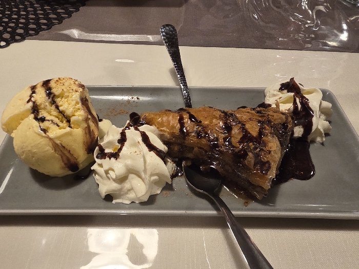 This image, taken in Osterhofen, Germany, depicts a dessert plate resting on a light-colored, textured tablecloth. On a rectangular grey plate, a single serving of dessert is presented. To the left, a large scoop of pale yellow ice cream is drizzled with dark brown chocolate syrup. In the center, a triangular piece of pastry, likely baklava, golden-brown and flaky, is heavily coated in dark brown chocolate syrup and lightly dusted with a reddish-brown powder, possibly cinnamon or cocoa. This pastry rests in a pool of dark liquid. Flanking the pastry are two distinct mounds of white whipped cream, each also adorned with dark brown chocolate syrup. Two metallic spoons are present; one small spoon is partially visible beneath the pastry on the left, and another spoon with a decorative handle is upright, inserted into the pastry. The background features a dark, intricately patterned placemat on the left and reflective surfaces, possibly glass or plastic, on the upper right. No individuals are visible, and no active actions or interactions are depicted. The environment appears to be an indoor dining setting.