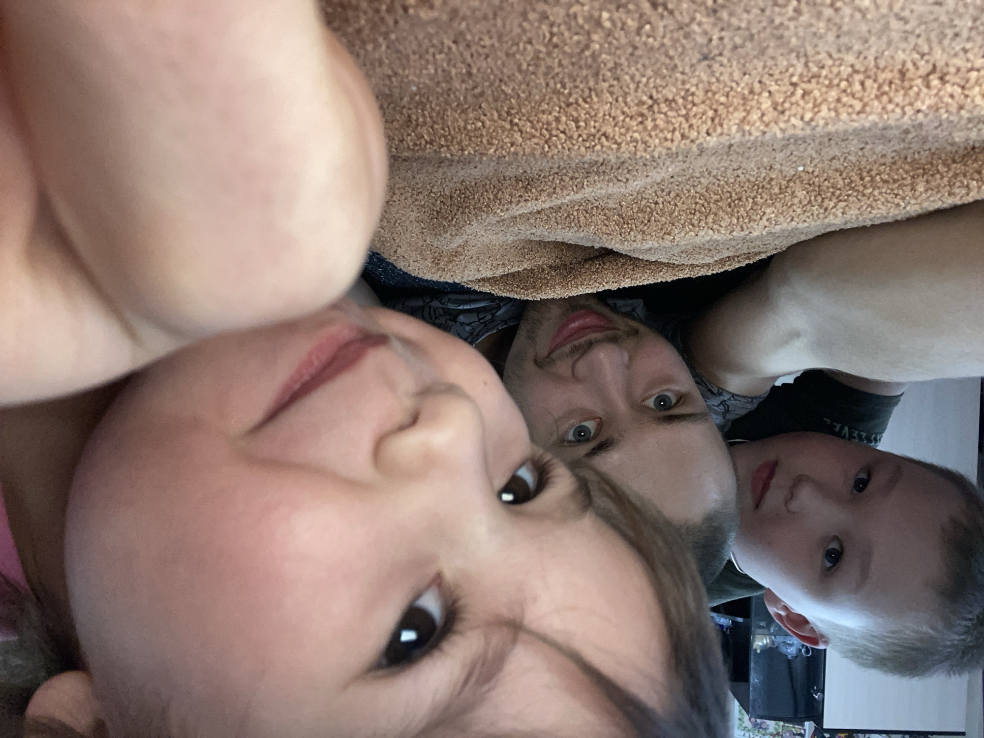 From an inverted perspective, the image shows three individuals and a textile object. A young child's face is in the foreground, occupying a large portion of the frame, partially obscured by hair. An adult male is positioned behind the child, his face visible. Another child is visible to the side of the man's face. The textile is above their heads. The location appears to be indoors, but the background is too blurry to discern specific details. There is no indication of Bely Klyuch, Russia.