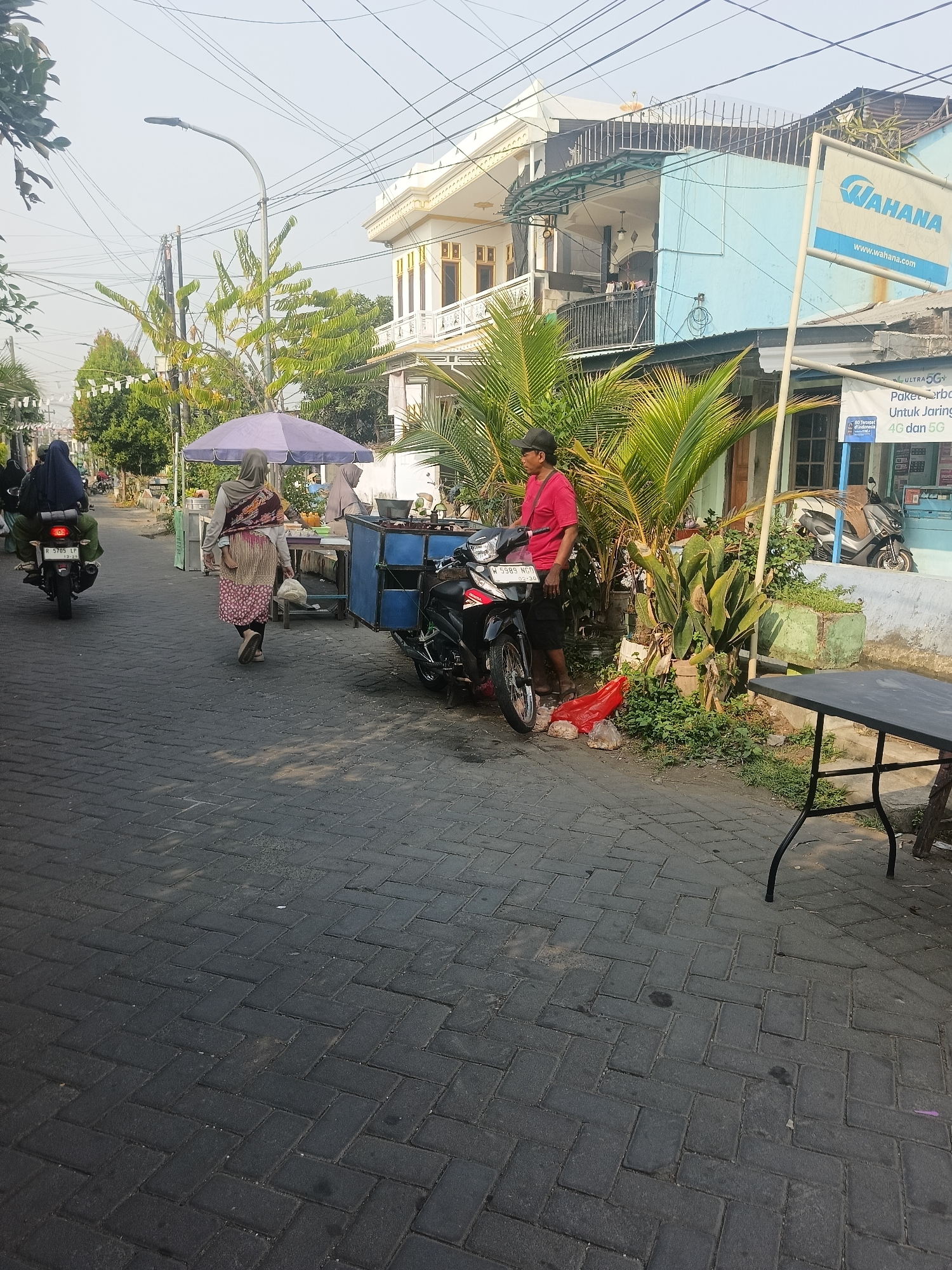 The image depicts a street scene in Gedangan, Indonesia. In the foreground, a paved road with interlocking bricks runs towards the viewer. To the right, a dark-colored table with metal legs stands on the pavement, casting a shadow. Beyond the table, a patch of green grass and vegetation is visible.
In the middle ground, a man wearing a red t-shirt and black shorts, and a black baseball cap, stands next to a motorcycle. He appears to be interacting with the motorcycle. The motorcycle is black and red, with the license plate reading "W 5985 NGO". To the left of the man and motorcycle, there is a blue cart with a metal frame, containing food items covered by a blue tarp. Further left, a woman in a hijab and a patterned skirt walks away from the camera, carrying a white plastic bag. Behind her, another woman in a black hijab is riding a motorcycle. A purple umbrella is set up near the blue cart, and a food stall is visible underneath it.
In the background, buildings line the street. On the right, a light blue building with white trim has a sign that reads "WAHANA" with a website address below it. Another sign with Indonesian text is also visible. A white building with ornate yellow trim stands further back. Trees and power lines are visible throughout the scene, indicating an urban or semi-urban environment. The overall lighting suggests it is daytime, possibly late afternoon due to the shadows.
