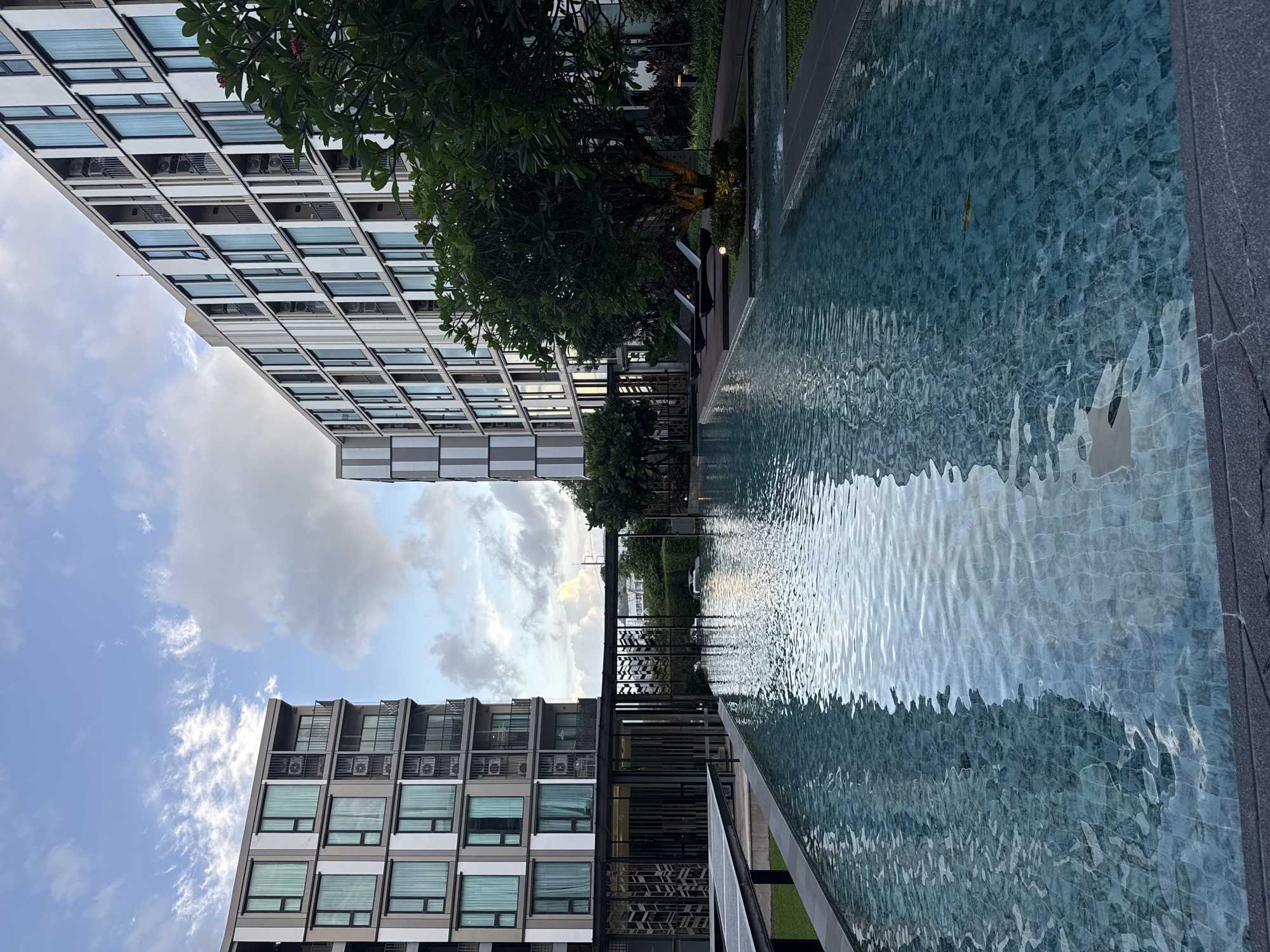 The image depicts an outdoor scene adjacent to a swimming pool. The swimming pool, tiled with small, blue-green squares, occupies the right side of the frame, its surface rippled by gentle waves. A dark, textured border runs along the edge of the pool.
On the left, a modern building with multiple floors is visible. The building's facade is characterized by repeating patterns of windows with light-colored blinds and individual air conditioning units. In the distance, another similar building can be seen, partially obscured by trees. Lush green trees and foliage are scattered throughout the scene, some with visible pink flowers, and also a large, gnarled tree with an exposed, twisted trunk. A dark metal fence with an intricate pattern runs between the building and the pool area.
The sky above is a mix of blue and white clouds, suggesting daytime, likely late afternoon due to the soft light reflecting on the water. The overall atmosphere appears serene and tranquil. No people are visible in the frame. The location is identified as Ratsada, Thailand. No text is visible in the image.
