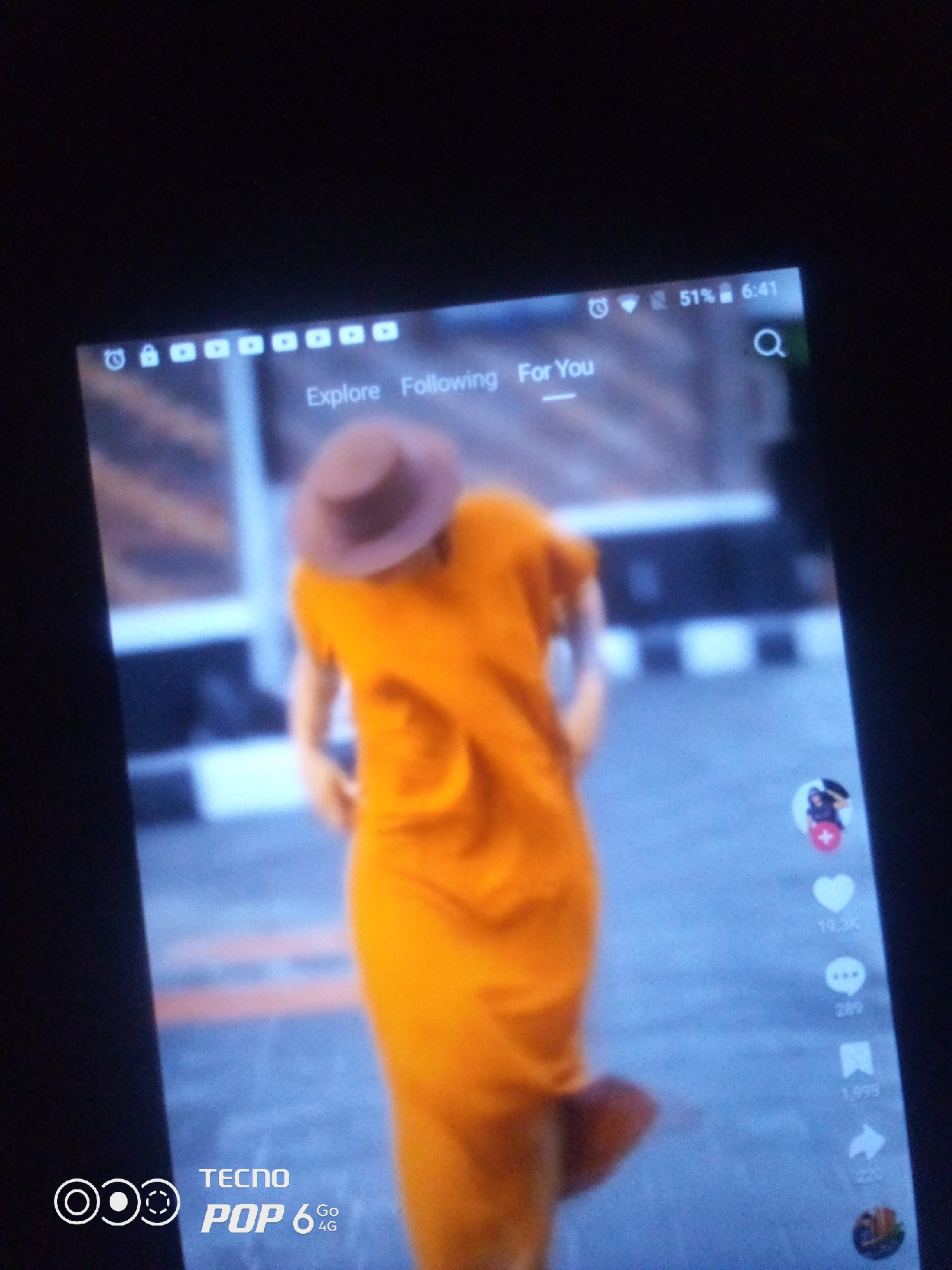 A vertically oriented, slightly blurry, close-up photo captures a person wearing an orange garment and a brown hat, viewed from behind. The person appears to be dancing or in motion. The background, a blurred city street, is likely in Howayi, Nigeria. The image displays a phone screen with a social media app interface, showing icons for likes, comments, shares, and user profiles. The phone's battery level is at 51% and the time is 6:41 AM. The phone model is indicated as TECNO POP 6 4G.