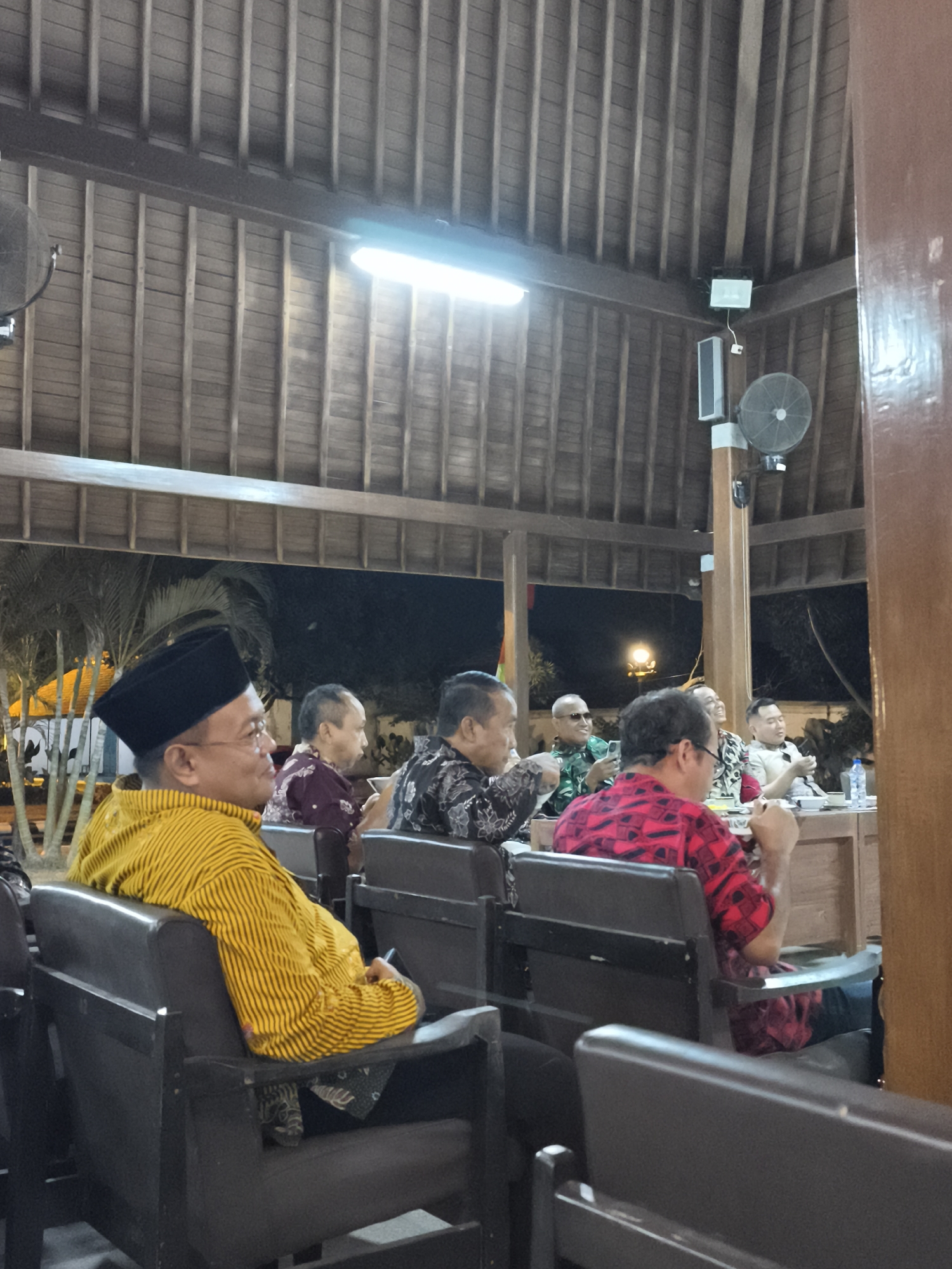 The image depicts a gathering of men seated in an outdoor pavilion, likely in Banyumas, Indonesia. The architecture features a wooden-slatted roof with exposed beams and a prominent brown wooden pillar on the right. Lighting comes from a long fluorescent tube fixture overhead and a spotlight further back, illuminating the space and the attendees.
In the foreground, a man in a yellow and black striped shirt and a black peci hat is seated in a dark chair, facing towards the right of the frame. He is wearing glasses and appears to be looking at something out of frame. To his right, other men are seated, some facing forward, others conversing. One man wears a maroon batik shirt, another a black and white patterned shirt.
In the middle ground, a group of men are seated around a wooden table. One man in a red and black patterned shirt is visible from the back, engaged in an interaction. Another man in a military-style green uniform and sunglasses is holding a phone. A man in a white shirt is also present at the table, along with others whose faces are not clearly discernible.
The background shows a dark exterior with palm trees visible, suggesting an evening or nighttime setting. A circular fan is mounted on a wooden pole in the middle ground. The overall atmosphere appears to be that of a formal or semi-formal meeting or event.