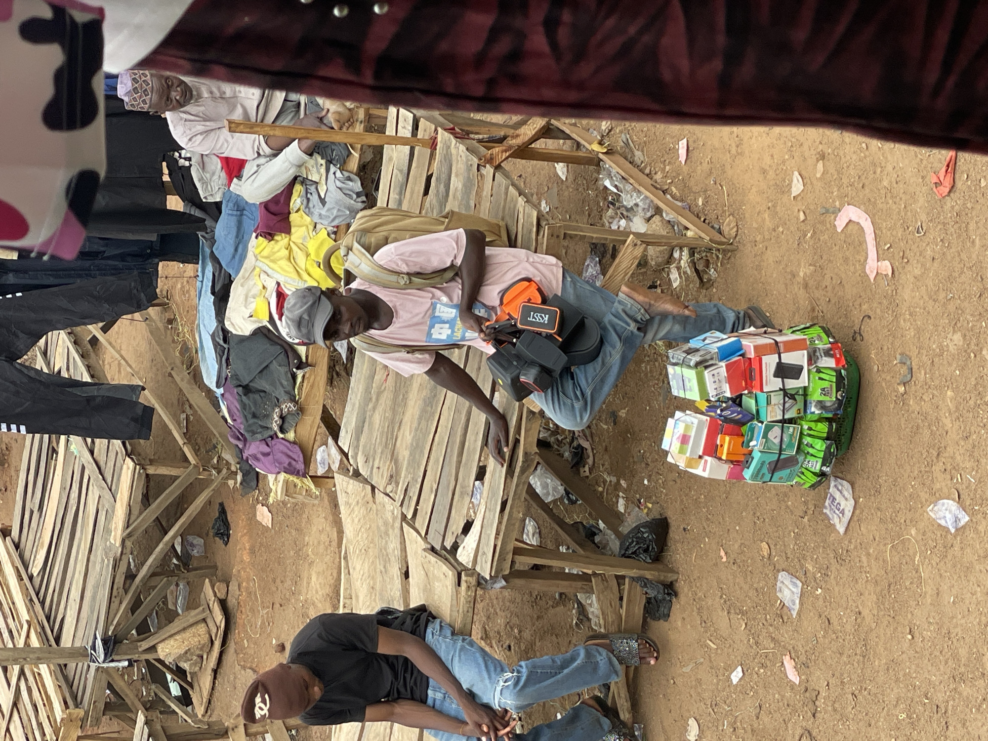 The image shows an outdoor marketplace scene in Bukuru, Nigeria. Two men are present. One man wears a pink t-shirt, jeans, a baseball cap, and has a backpack. He is sitting on a rudimentary wooden structure and appears to be operating a small electronics business, with a pile of packaged electronic items (possibly mobile phones and accessories) beside him on the ground. The other man, wearing a black t-shirt, jeans, and a brown cap, is sitting on the ground near the wooden structure. Clothes are hanging from the structure. The ground is dirt, with trash and debris scattered around. The market appears to be in a low-income area.