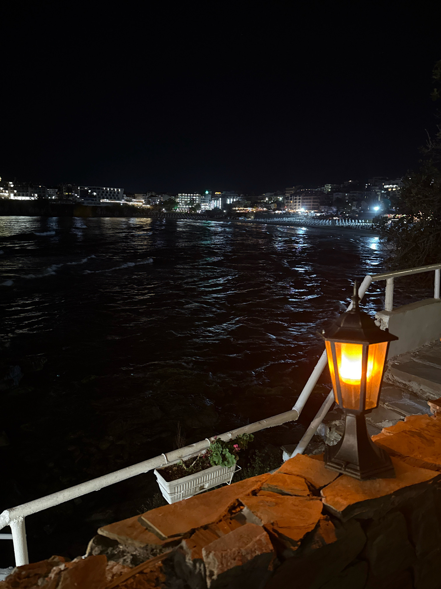 The image depicts a nighttime scene at the seaside in Sozopol, Bulgaria. In the foreground, a lit lantern with an orange glow illuminates a stone wall and a white railing. Beyond the railing, the dark water of the sea is visible, with small waves reflecting the lights from the distant town. The town, situated along the coastline, is dotted with numerous buildings illuminated by lights, creating a vibrant urban backdrop against the dark sky. The overall atmosphere is serene and inviting, suggesting a peaceful evening by the sea.