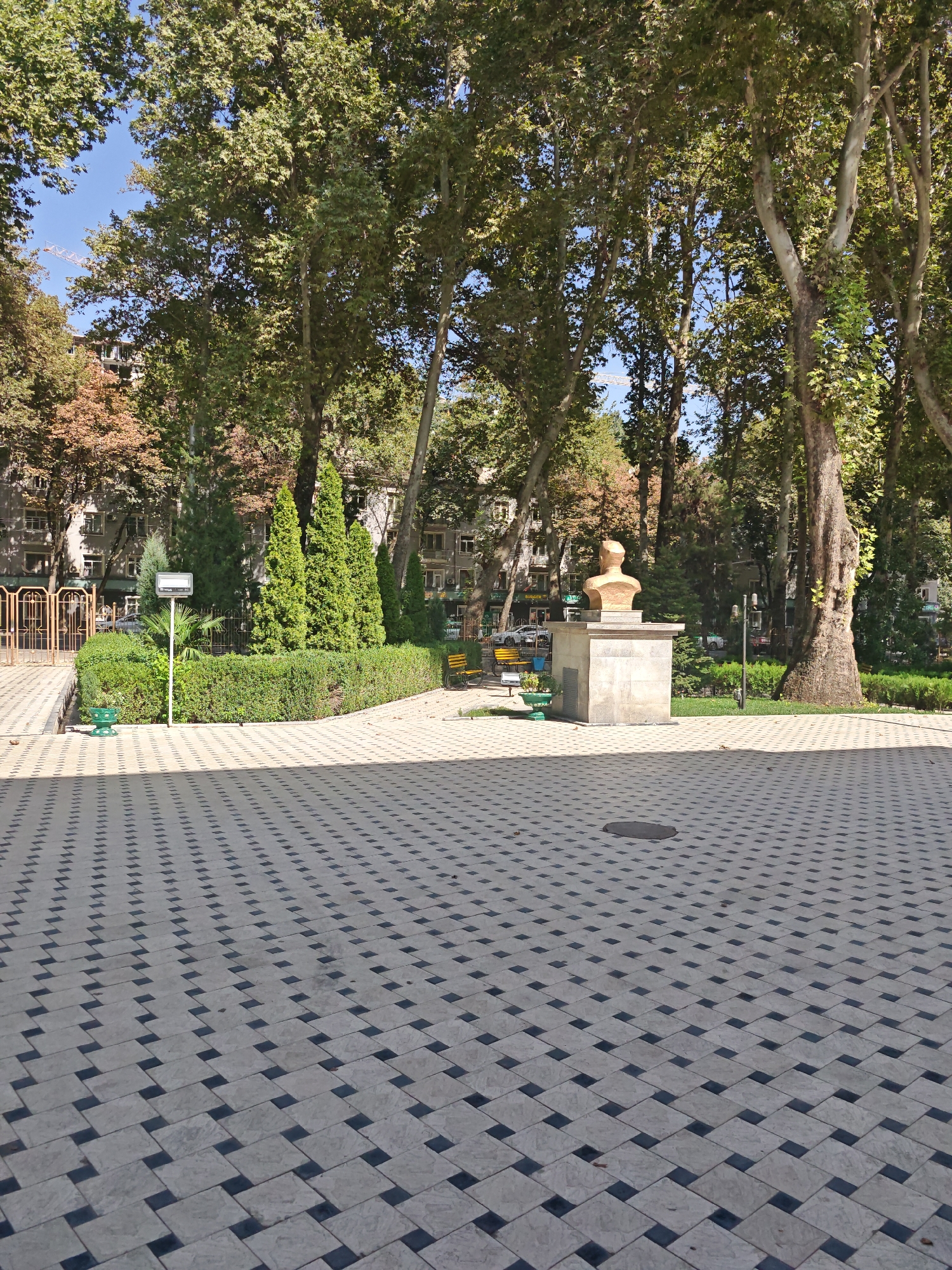 The image shows an outdoor plaza with a distinctive patterned paving. In the foreground, the paving consists of light-colored square tiles arranged in a diagonal, overlapping pattern with darker square tiles interspersed. The paving extends into the midground, where it meets a lawn area.
To the left of the lawn, there is a yellow metal fence with decorative elements and a pathway leading away from it. A green planter is situated near the fence. A white signpost with a rectangular sign is also visible.
In the midground, behind the lawn, is a sculpture. It consists of a bust of a person, made of a golden-bronze material, placed on top of a stone pedestal. The pedestal is rectangular and appears to be made of light-colored stone. Behind the sculpture, there are various trees, including tall, mature trees with broad leaves and some evergreen trees.
Further in the background, residential buildings with multiple windows are visible behind the trees. The sky is clear and blue. The scene is set during the daytime, with sunlight casting shadows on the paving, indicating a bright, sunny day. The overall atmosphere is one of a public park or square.
