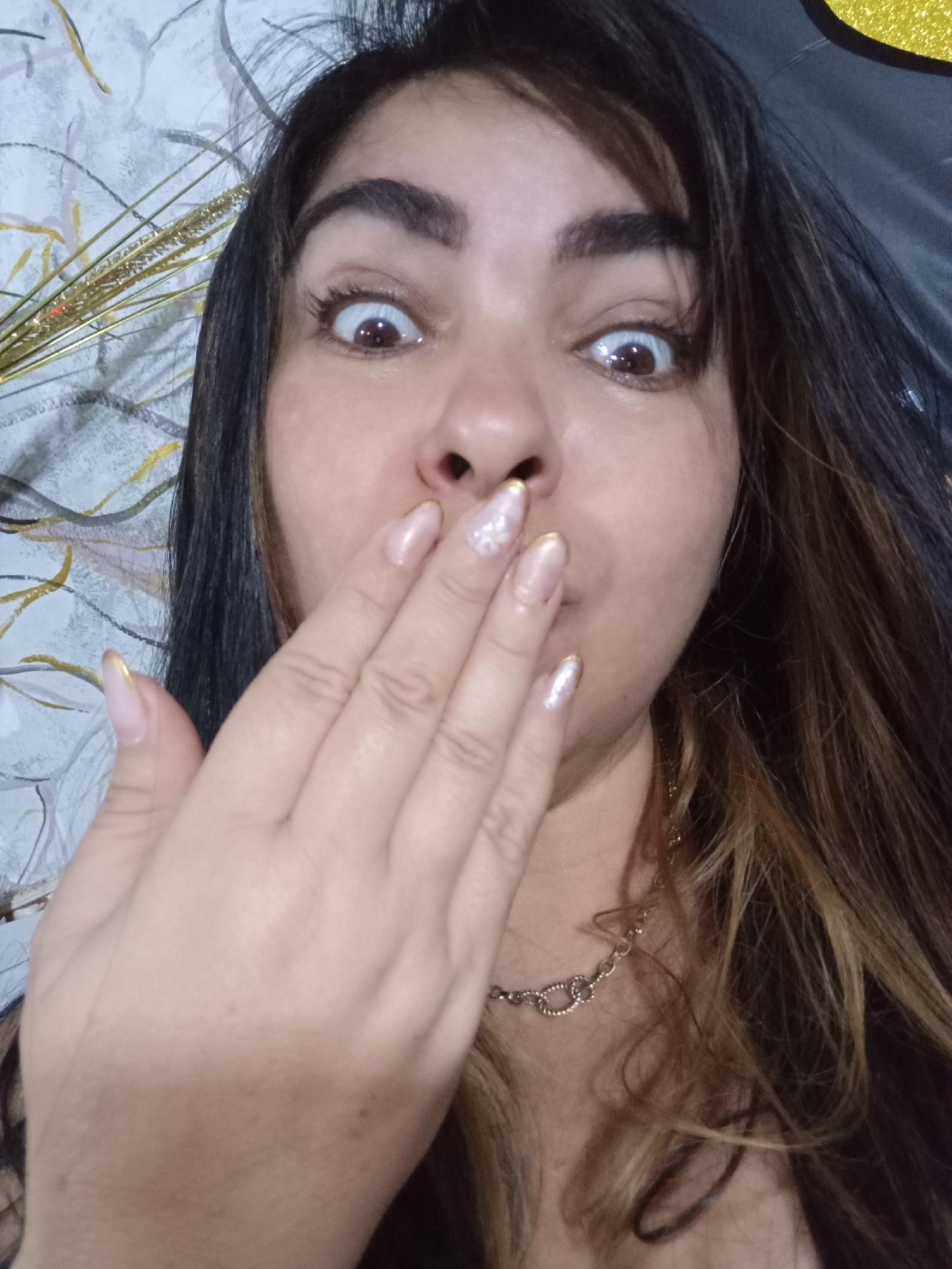 The image presents a close-up, chest-up indoor portrait of a female individual with dark, long hair featuring lighter brown highlights and bangs. Her eyes are wide open, conveying an exaggerated expression of surprise or shock, with her eyebrows slightly raised. The individual's right hand is positioned in front of her mouth, with her fingers partially covering her lips and nose. Her fingernails are manicured with an elongated oval shape, painted a light, translucent color with distinct gold tips; one nail on the index finger appears to have additional decorative embellishments. A silver-toned, linked chain necklace is visible around her neck, resting against her chest. The background consists of a light-colored wall with a subtle, abstract linear pattern in muted tones of grey and gold. To the left of her head, shiny, thin gold decorative strands are visible. Towards the upper right, a dark, possibly glittery, curved object is present. The lighting suggests an artificial indoor setting. The specific location within Juan Griego, Venezuela, cannot be determined from the visual information provided in the image.