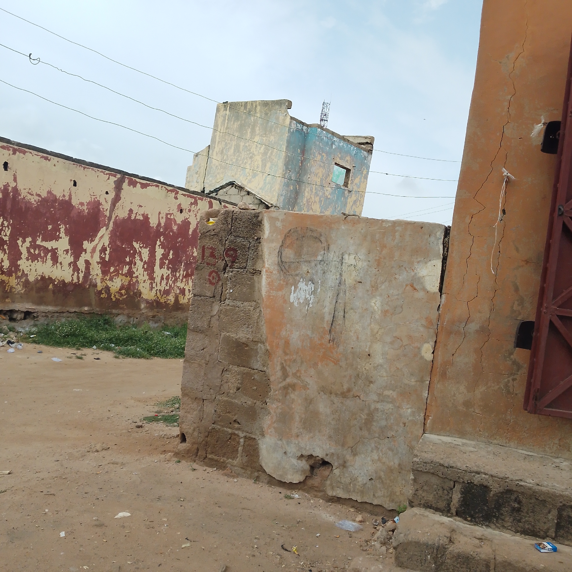 The image depicts a street scene in Pantame, Nigeria, characterized by dilapidated buildings and an unpaved ground. In the foreground, a rough, weathered concrete pillar stands, its surface marked with graffiti, including what appears to be a circular drawing and some vertical lines. To the left of this pillar, a section of a brick wall is visible, also showing signs of wear and disrepair, with red markings on it.
Behind the pillar, a long, low building with a faded red and cream exterior stretches across the midground. The paint on this building is peeling extensively, revealing the underlying surface. Further back, a more imposing, multi-story structure dominates the background. This building is leaning precariously, with sections of its light-colored facade covered in patches of blue and yellow paint, and a visible square opening where a window might have been. A communication tower or antenna is perched atop this leaning structure.
The ground in the scene is sandy and dusty, with patches of sparse green grass growing near the base of the buildings. Debris, including plastic wrappers and other small litter, is scattered across the ground. A tall, orange-colored wall with significant vertical cracks runs along the right side of the image, partially obscuring a dark metal gate or structure. Overhead, electrical wires are strung across the sky, which is overcast and pale. The overall impression is one of neglect and urban decay. No people are visible in the frame.