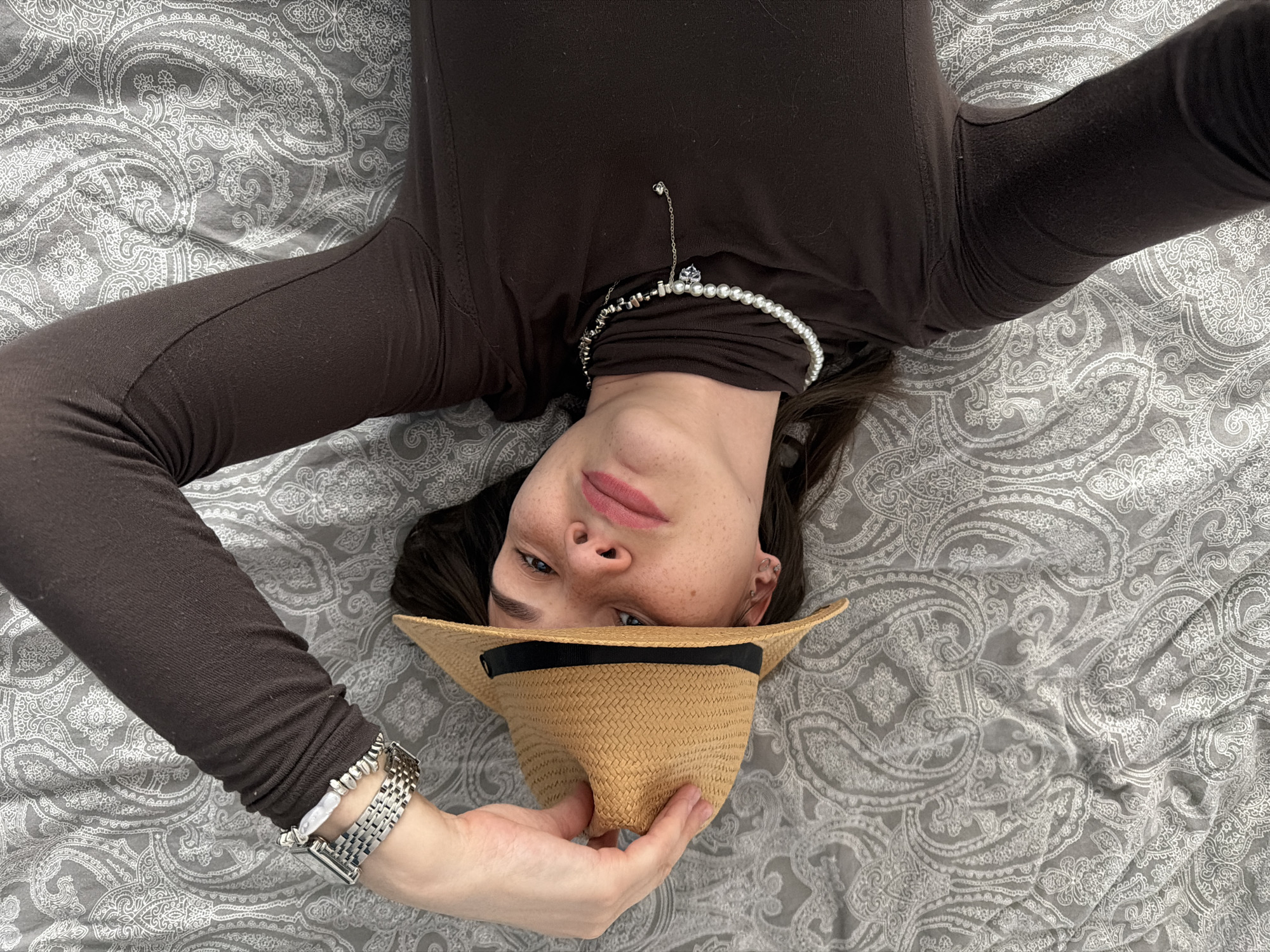 An overhead shot captures a person, appearing to be an adult female, lying on their back, looking upwards towards the camera. The individual is positioned inverted in the frame, with their head at the bottom. They have dark hair, blue eyes, and noticeable freckles across their nose and cheeks. Their lips are painted a soft pink.
The person is wearing a long-sleeved, dark brown top. Adorning their neck are two necklaces: one made of pearls and another silver-toned chain with small beads. On their left wrist, they wear a silver-toned wristwatch with a metal link band, alongside another bracelet composed of light-colored beads.
Their left hand is holding a light brown straw hat with a black band, positioned near their face, slightly obscuring part of their chin and mouth. The pose appears relaxed and casual, with a neutral to slightly smirking expression.
The setting is an indoor space, likely a bed, covered with a grey and white patterned fabric, featuring an intricate paisley design. The lighting is soft and even, suggesting an indoor environment without direct harsh sunlight. There is no visible text in the image. The location context provided is Chaikovsky, Russia.