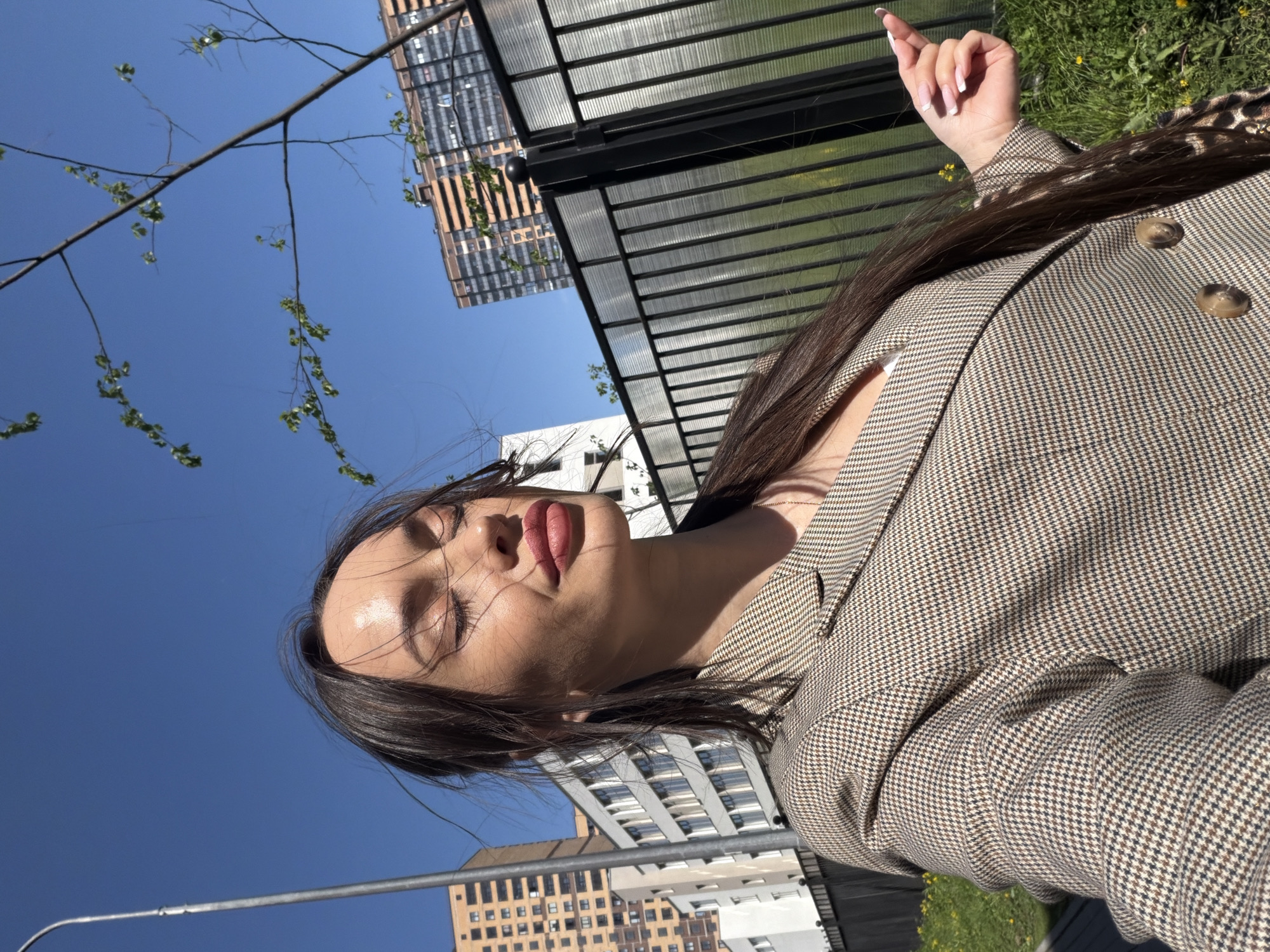 A photo taken in the city of Unknown, Russia, presents a woman in a brown patterned blazer with two visible buttons, standing near a black metal fence. She is holding up her right index finger, suggesting she is in the midst of communication or pointing at something out of view. The photo appears to be taken from a low angle, revealing the sky and several buildings in the background. A thin tree branch with budding leaves is also visible in the top-left corner. The environment includes a patch of grass and a structure with a translucent covering.