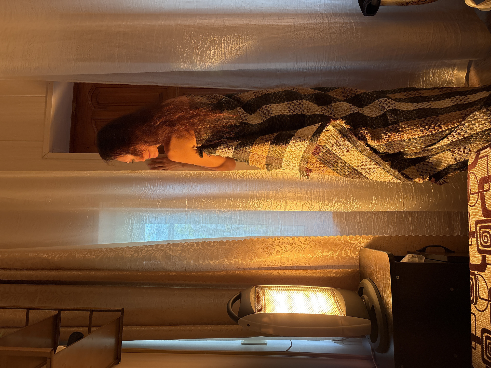 A woman lies in bed covered with a patterned blanket. The scene is illuminated by the glow of a space heater in the foreground. A window is partially visible behind the bed, with light filtering through curtains. A wooden doorframe is above the woman's head. A dark brown cabinet is to the right, and a wooden structure with shelves is to the left. The location is speculated to be in Gelendzhik, Russia.
