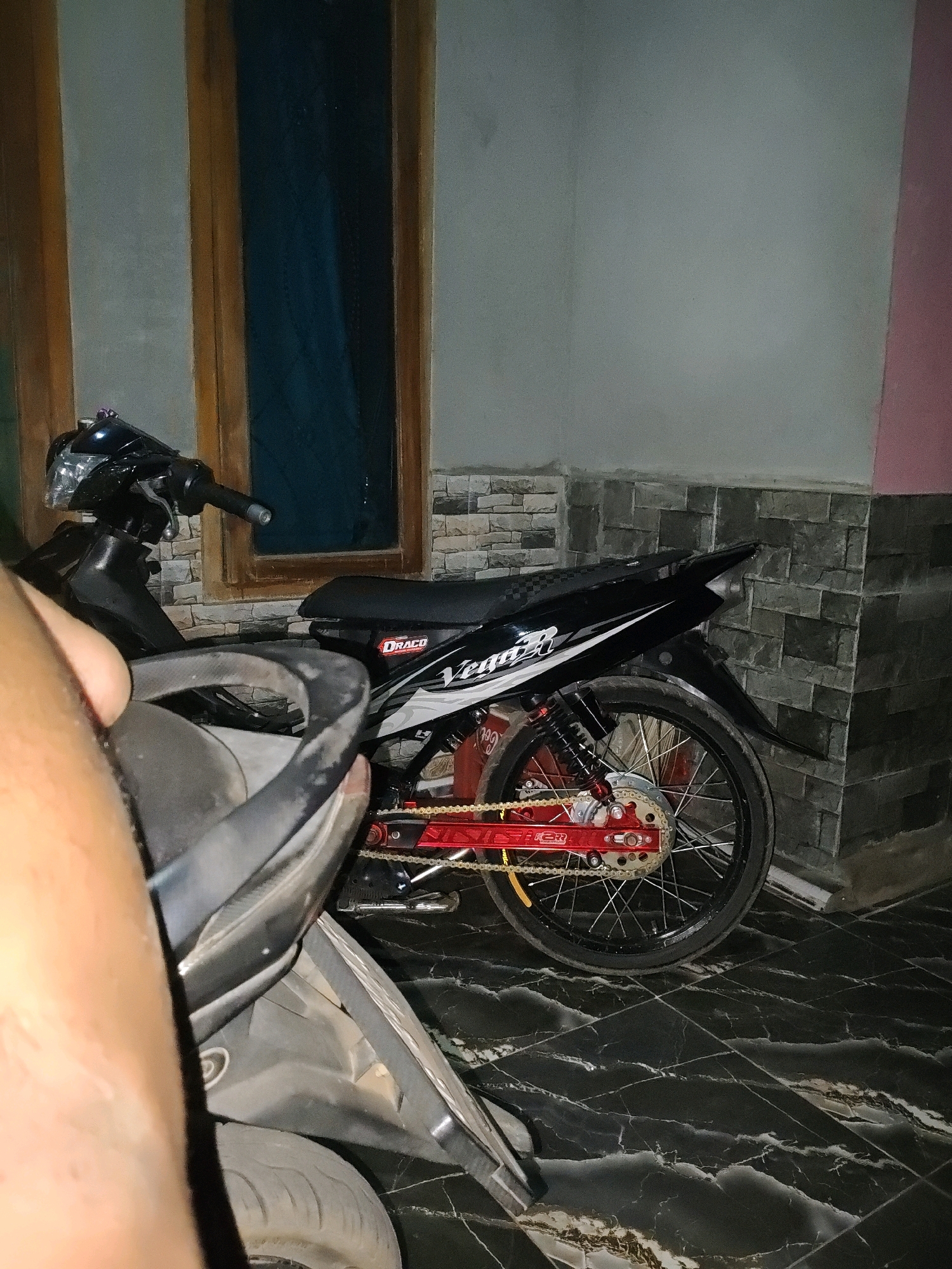 The image depicts a street view in Karang Sari, Indonesia, showing two motorcycles parked in an outdoor area. One motorcycle, a black scooter, is partially obscured by a person's arm in the foreground. The second motorcycle, also black, is a sport bike with red accents on its chain and rear suspension. It has a "DRACO" sticker on its side. The motorcycles are parked on a dark, marble-patterned tiled floor. Behind the motorcycles, there is a wall with grey stone tile covering the lower portion and a light grey painted upper portion. To the left, a wooden-framed window with blue curtains is visible. The lighting suggests it is either dusk or nighttime.