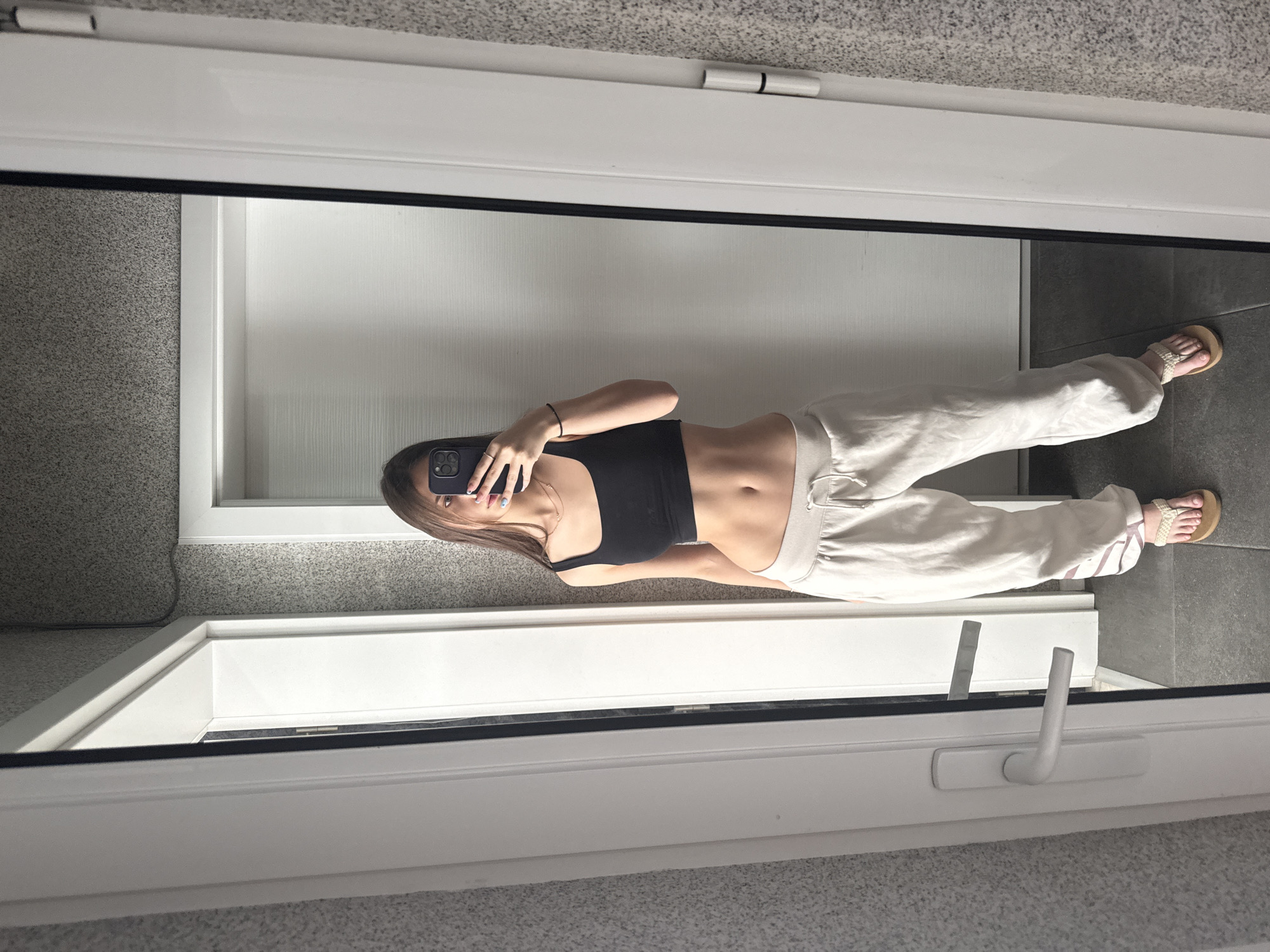The image captures a young woman taking a mirror selfie in an indoor setting, identified as being in the city of Sofia, Bulgaria. The central subject is a female with long, straight dark brown hair, positioned directly in front of a large, rectangular mirror. Her face is mostly obscured by a dark-cased smartphone, likely an iPhone, which she holds up with her right hand to take the photo. Her left arm is visible, bent at the elbow.
The woman is wearing a black, fitted sleeveless crop top that reveals her midriff. Her lower body is clad in light beige or off-white sweatpants with a visible drawstring at the waist. She is wearing light-colored thong sandals with a woven strap detail, revealing manicured toenails. Accessories include a delicate, layered necklace with two chains around her neck, a ring on her right hand, and a black bracelet on her left wrist. Her right hand fingers display light blue nail polish.
The environment, visible both directly and as a reflection in the mirror, features walls covered in a light grey, speckled texture, resembling wallpaper or textured paint. The floor is composed of large, dark grey rectangular tiles. Two white interior doors with matching white frames are prominent. The door on the viewer's left (reflected on the right side of the woman) is equipped with a modern white handle. The door on the viewer's right (reflected on the left side of the woman) shows a visible silver hinge. The mirror itself has a thin, dark border. The scene is illuminated by uniform ambient indoor lighting.