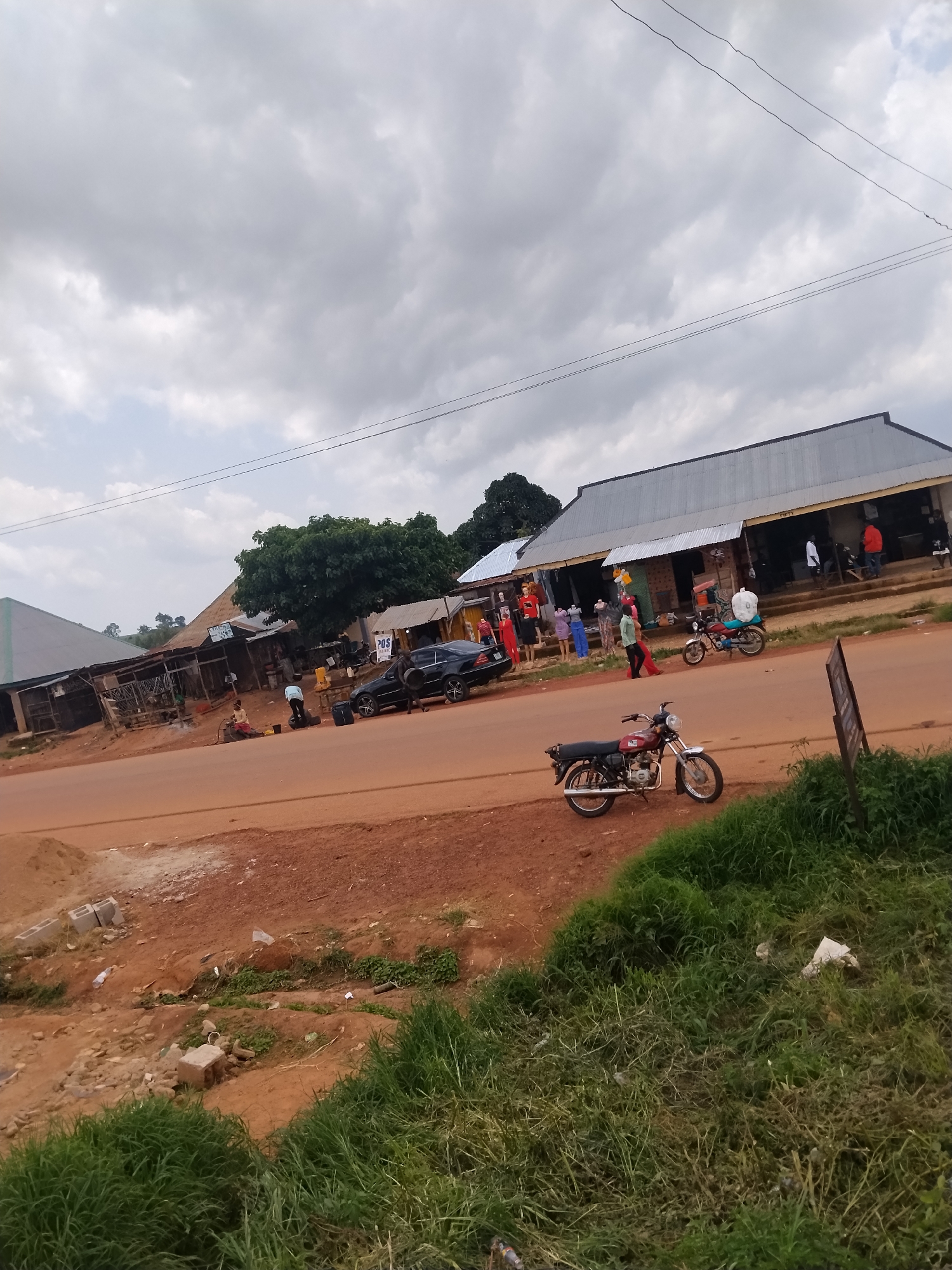 The image depicts a roadside scene in Durumi, Nigeria, under a cloudy sky. On the right side of the red dirt road, a row of shops with corrugated metal roofs lines the street. Several people are visible near the shops, some interacting with each other, while others appear to be customers or vendors. A black sedan is parked on the side of the road, and a red motorcycle is parked in front of one of the shops. Further down the street, another motorcycle is visible. The foreground shows a grassy embankment with some scattered debris. The overall environment appears to be a commercial or residential area with unpaved roads and modest structures.