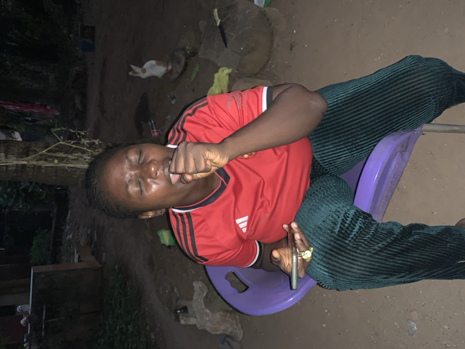 A woman sits on a purple plastic chair outdoors. She wears a red shirt with black stripes and dark green corduroy pants. She has a cell phone in her right hand, which is adorned with multiple rings. Her left hand is clenched into a fist near her mouth. Her eyes are closed, and she appears to be in pain or discomfort. The background is dark with some foliage and other objects visible.