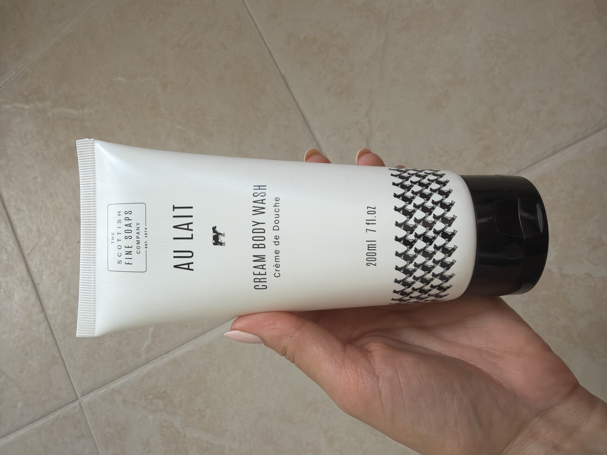 A hand holds a white tube of "Au Lait" cream body wash by The Scottish Fine Soaps Company. The tube features a black cap and a black and white pattern of cows near the bottom. The background is a tiled floor. The location is Pazardzhik, Bulgaria.