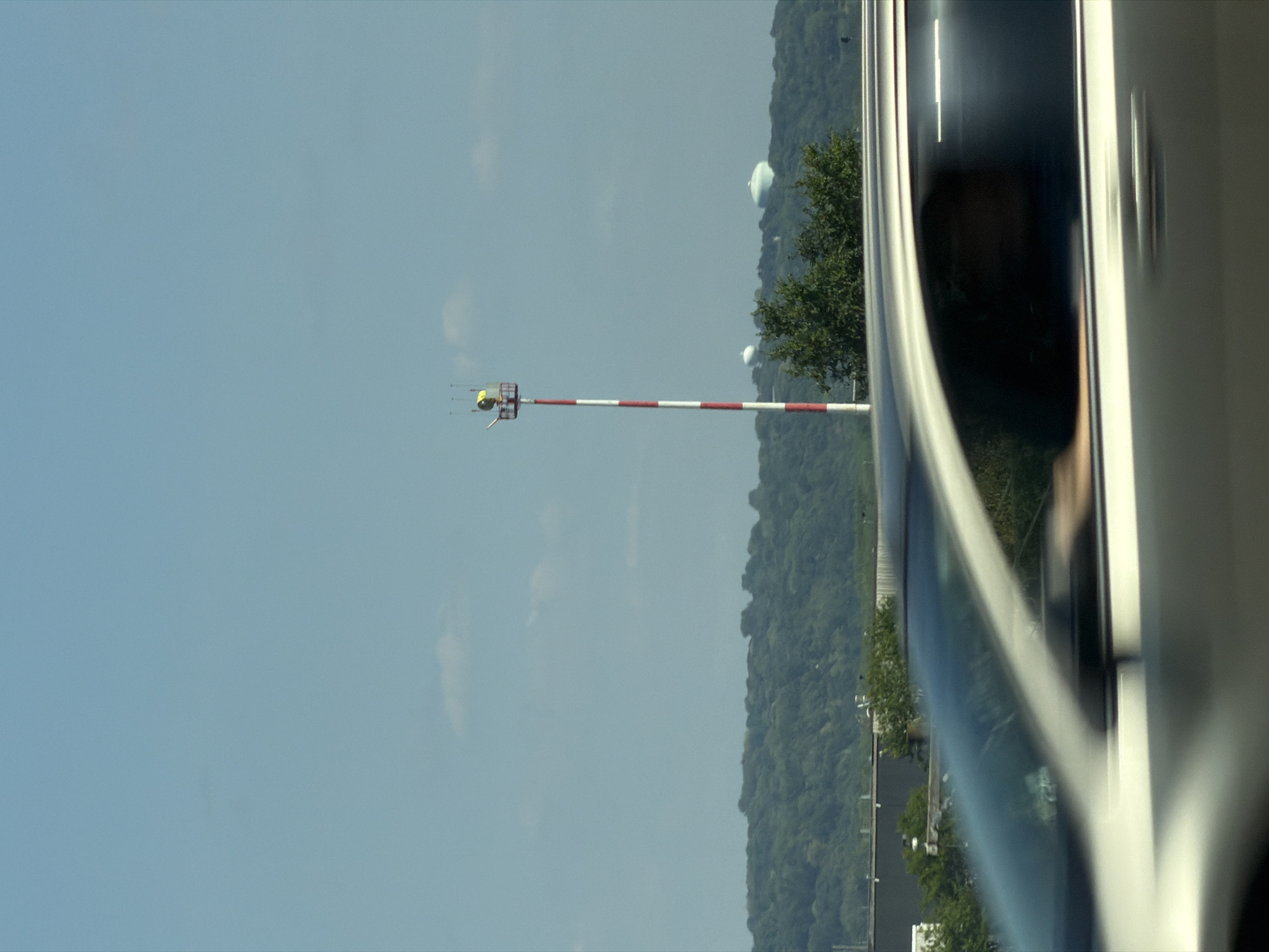 A striped red and white pole with a yellow light at the top stands against a blue sky. In the background, a line of green trees stretches across the horizon, with two rounded white structures visible above the trees. In the foreground, the side of a silver car is blurred due to motion, with a hint of a person's reflection in the window. This image appears to be taken from a vehicle traveling on I-94 Westbound near the Highway 280 interchange in Saint Paul, Minnesota, United States of America.