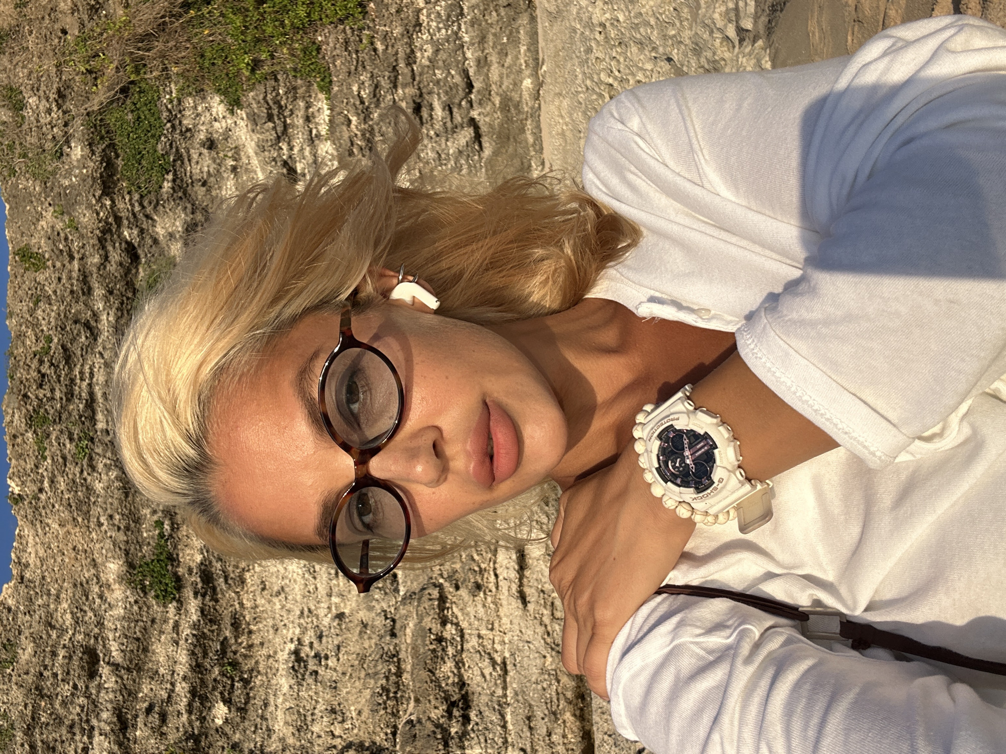 In Kuta Selatan, Indonesia, a woman with blonde hair and fair skin is seen in a selfie. She is wearing a white collared shirt, oval-shaped glasses with dark frames, and a white wristwatch with a dark face. She has on white wireless earbuds in her left ear. She is standing in front of a rocky cliff face, which appears to be made of sandstone, with some green vegetation growing on it. The lighting suggests it is either sunrise or sunset, with a warm glow illuminating the scene. The woman's left hand is resting on her left shoulder. The background is slightly out of focus, with a hint of blue sky at the top.