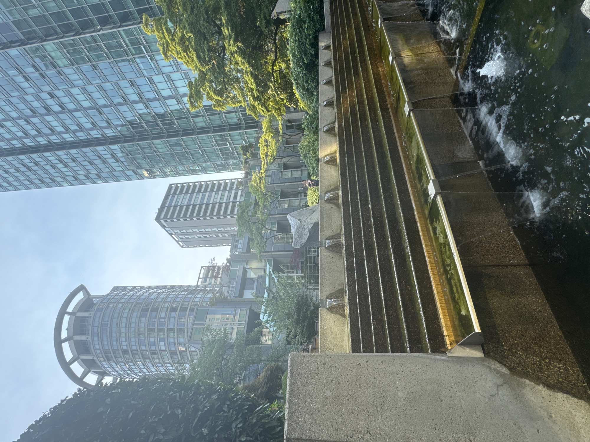 The image depicts a tiered water feature in a contemporary urban setting, likely located in Vancouver, Canada. The foreground is dominated by concrete steps and cascading water, with a dark, reflective pool at the bottom. The water feature is flanked by concrete structures and lush green foliage. In the background, several modern skyscrapers rise against a clear blue sky. One prominent circular building with a distinctive roof structure is visible to the left, while a taller, rectangular glass-facade building stands to the right. The scene suggests a well-maintained public space within a city. A person is visible in the mid-ground, standing near the water feature.