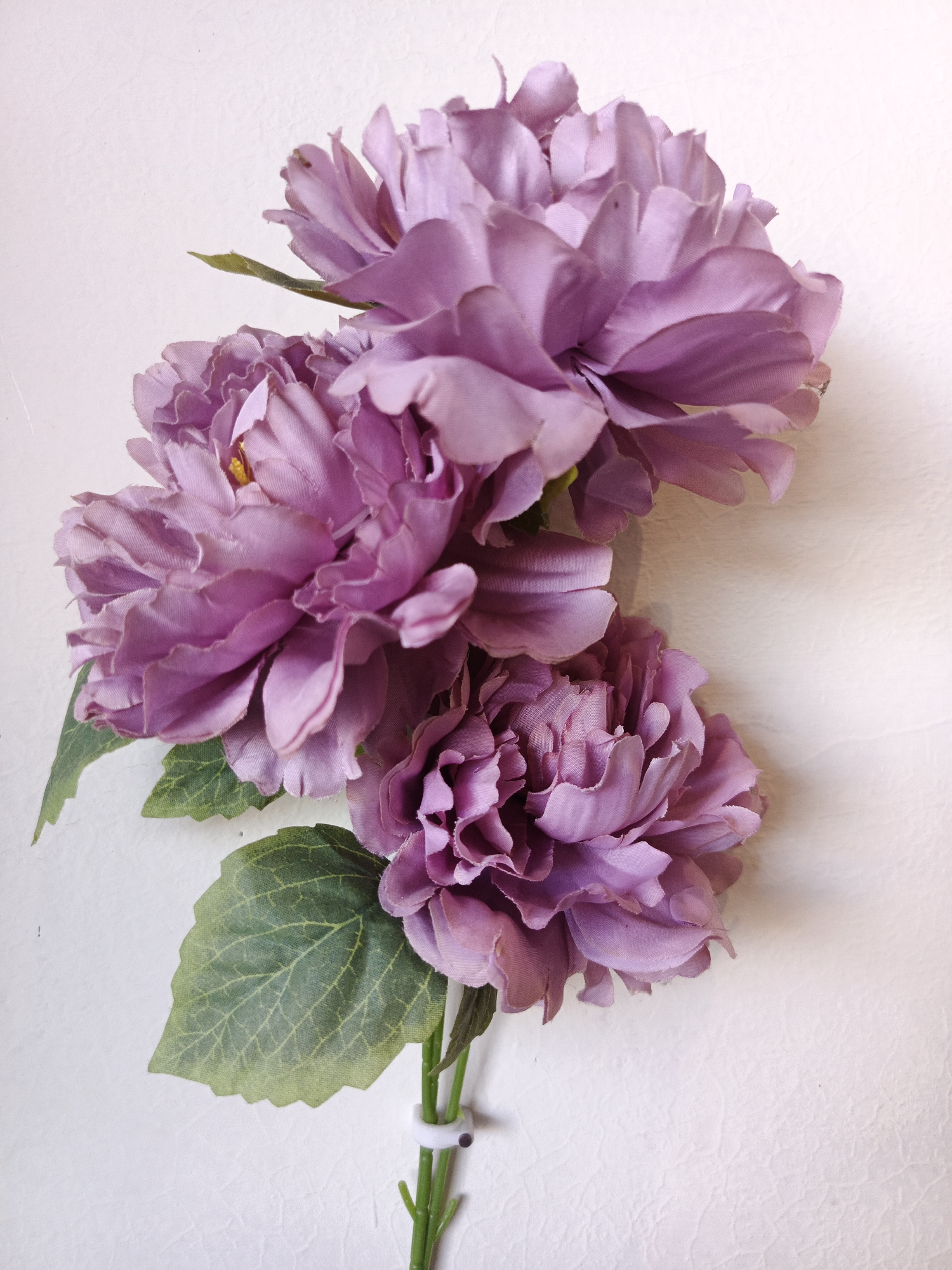The media file is a close-up, eye-level shot of artificial purple peonies against a textured, off-white background. The lighting is bright and diffused, suggesting an indoor setting with no distinct shadows. There is no discernible time of day or weather indicated. The image focuses on a bouquet of three lush, multi-layered peonies, with visible green leaves. The flowers are detailed, showing the fabric texture and the layered arrangement of their petals. A small white zip tie is visible at the bottom of the stems, holding them together. There is no text present in the image. The setting appears to be a still life composition.