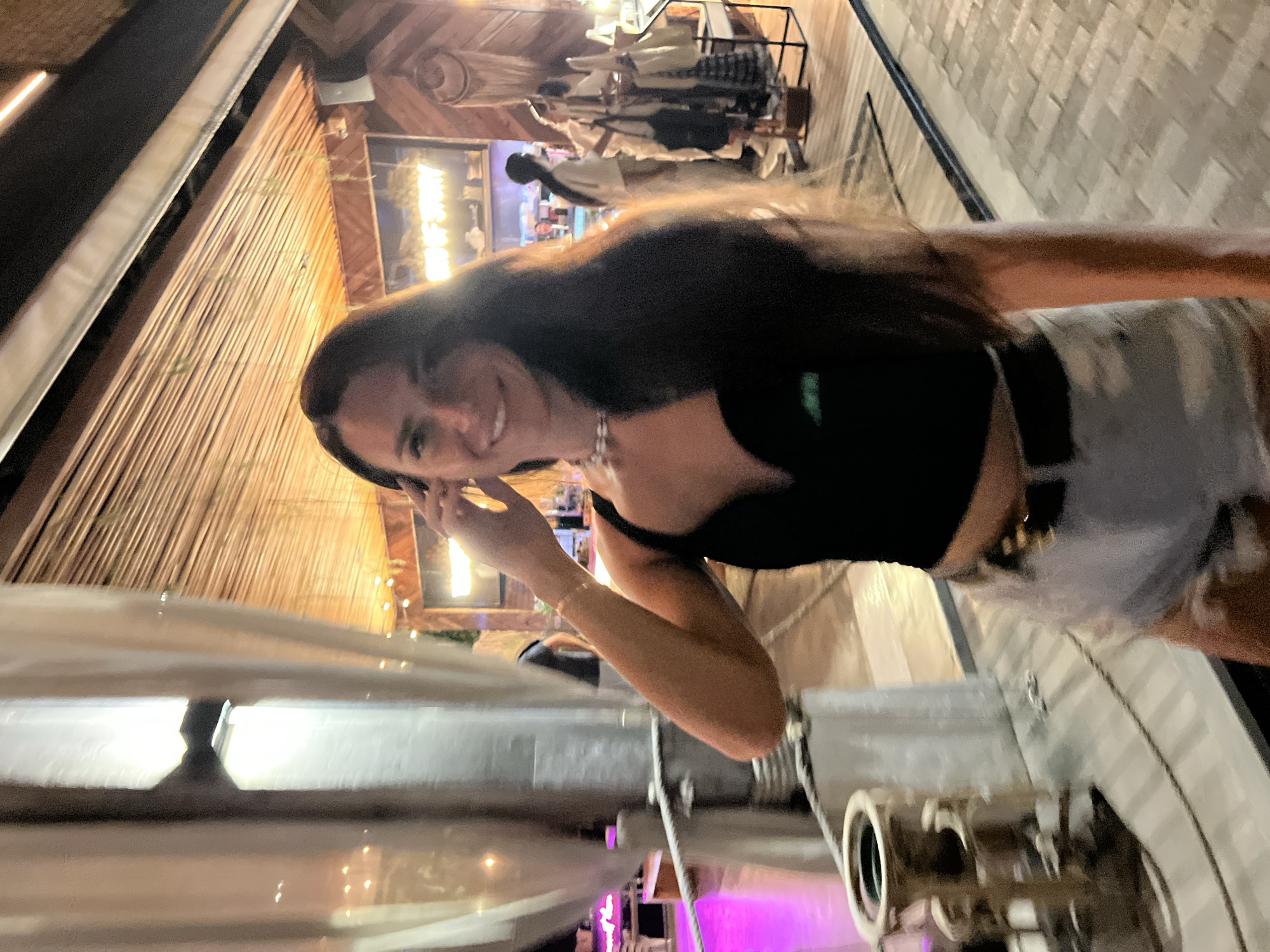 In Baan Bo Phut, Thailand, a young woman with long, dark hair stands in what appears to be an outdoor restaurant or market setting at night. She is wearing a black crop top and denim shorts. Her right hand is raised to her head, and she is smiling at the camera. Behind her, there are thatched ceilings, wooden accents, and displays of clothing and accessories. The scene is illuminated by warm artificial lights, with a hint of pink lighting visible in the background. The ground appears to be paved with brick-like stones.