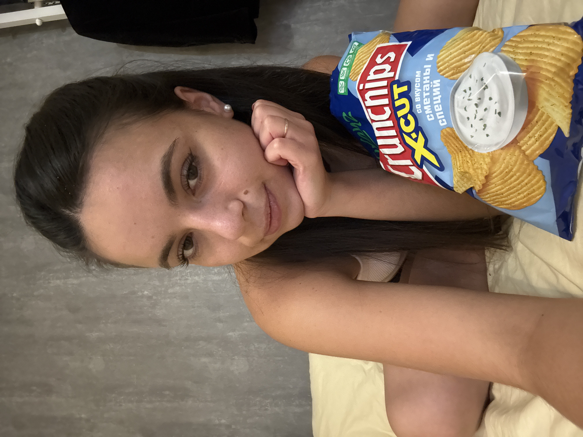A young woman with long, dark hair is lying down, holding a bag of Crunchips X-Cut potato chips. Her right hand rests on her cheek, and she is wearing a ring and a pearl earring. The bag of chips is blue and features images of ridged potato chips and a bowl of dip. Text on the bag includes "Crunchips X-cut" and Russian text that translates to "with sour cream and herbs flavor." The woman is wearing a light-colored top. The background appears to be a bed with a pale yellow sheet, and a wall with a textured gray surface. The lighting suggests it might be indoors during the day. The woman's expression is relaxed and appears to be a selfie.