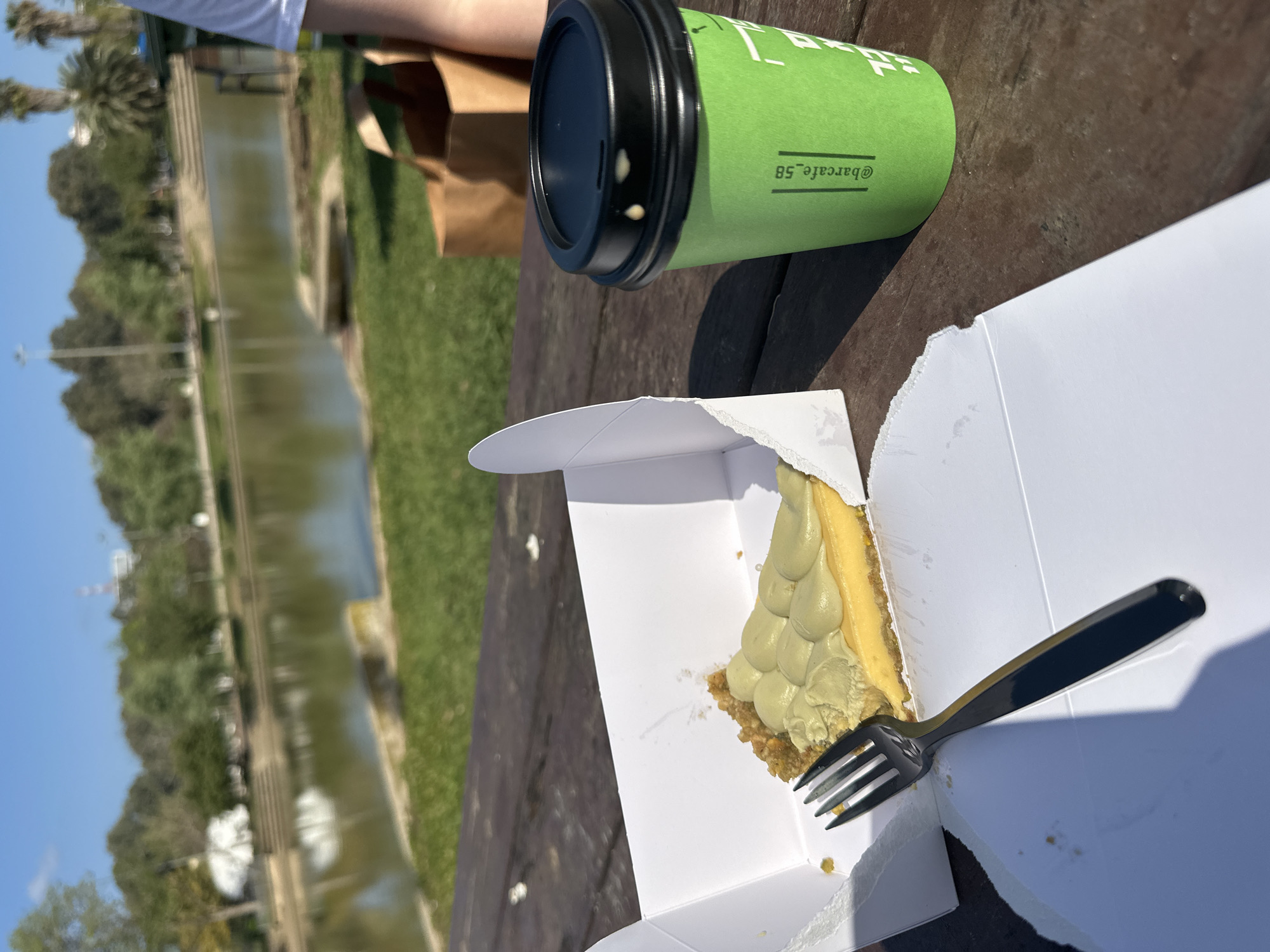 This image captures a relaxed outdoor setting, likely in a park or by a canal, under bright daylight. The scene is viewed from a slightly elevated perspective, looking across a wooden table towards a body of water.
In the foreground, on the right side of the wooden table, are several items. A vibrant green disposable coffee cup with a black lid is prominently featured, with two small drops of liquid visible near the lid's opening. Below the cup, a white cardboard box contains a slice of what appears to be a creamy tart or pie, possibly key lime or pistachio, indicated by its light green, piped topping and a crumbly crust. A black fork rests inside the box next to the dessert, suggesting it is either about to be eaten or has just been sampled. Partially visible in the upper right corner is the arm of a person wearing a white short-sleeved top, suggesting they are seated at the table. A brown paper bag is also partly visible behind the coffee cup on the table.
The background, on the left side of the image, features a calm, greenish-brown body of water, resembling a canal or a narrow river, with green grassy banks on either side. Lush trees, including palm trees, line the banks under a clear, bright blue sky. The overall impression is one of leisure and enjoyment of refreshments outdoors on a sunny day.
A notable detail is the visible text on the green coffee cup: "@barcafe_58", likely a social media handle for the establishment. Given the location context of Tel-Aviv, Israel, this setting could be a scenic park like Hayarkon Park. The strong shadows indicate a clear, sunny day, likely morning or early afternoon.