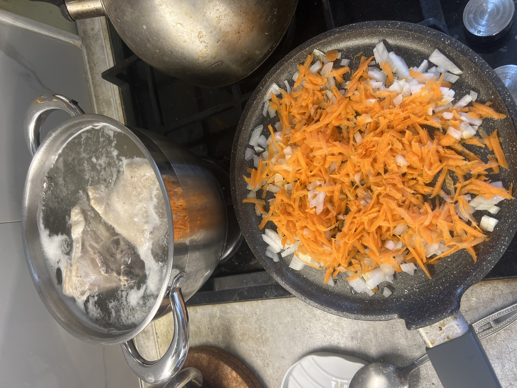 A close-up, overhead shot captures two cooking activities on a stovetop. In the foreground, a dark frying pan is filled with a mixture of shredded carrots and diced onions, ready for sautéing. In the background, a large metal pot is filled with simmering liquid and what appears to be pieces of meat or bone, suggesting a broth is being prepared. To the right, a silver kettle rests on the stovetop. The scene is set in a kitchen environment, likely within a residential apartment. No people are visible in the frame. The image quality is sharp, providing clear detail of the food ingredients and cookware. No specific location information is available within the image itself to pinpoint it to Volgograd, Russia.