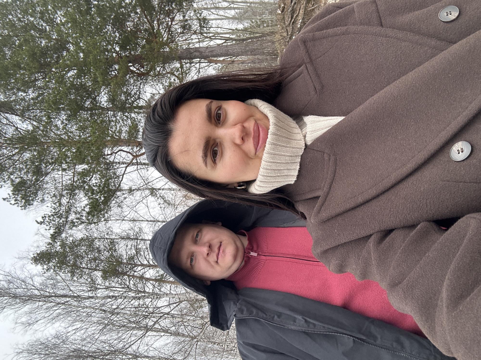 The image, rotated 90 degrees clockwise, captures a selfie of a woman and a man in a wooded outdoor setting, likely in Chaikovsky, Russia. The scene suggests a cold season, such as winter or early spring.
In the foreground, a woman with long, dark hair and a warm smile looks directly at the camera. She is wearing a cream-colored turtleneck sweater and a dark brown, possibly wool, coat with visible buttons. To her left and slightly behind her, a man with light hair and blue eyes looks towards the camera with a calm, neutral expression. He is wearing a dark grey or black hooded jacket over a pink or magenta zippered sweater, with the hood up.
The background consists of a forest or park, featuring a mix of bare deciduous trees and green coniferous trees under an overcast, bright sky. Both individuals are dressed in warm attire suitable for chilly weather. There is no visible text in the image.