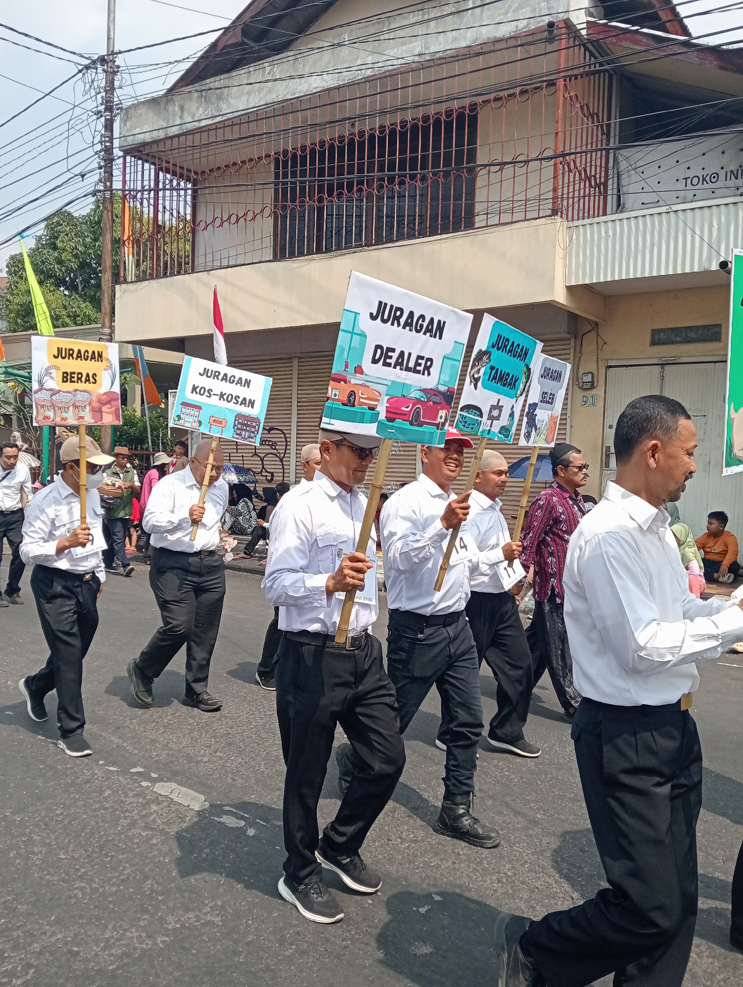 The image captures a street procession in Bintoro, Indonesia, under a clear sky. A group of men, mostly dressed in white shirts and black trousers, are walking in formation. They are carrying signs on wooden poles. The signs feature text in Indonesian, such as "JURAGAN BERAS" (Rice Merchant), "JURAGAN KOS-KOSAN" (Boarding House Merchant), "JURAGAN DEALER" (Dealer Merchant), "JURAGAN TAMBAK" (Pond Merchant), and "JURAGAN SELEB" (Celebrity Merchant). Some of the signs also have illustrations relevant to their respective titles.
Behind the procession, buildings line the street. One building has a prominent balcony with a metal grill. Another building has signage that appears to say "TOKO INDO". The street itself is paved and shows signs of wear, with faint white lines indicating a road marking. A few onlookers are visible in the background, some sitting on the side of the road. The overall atmosphere suggests a local event or parade.