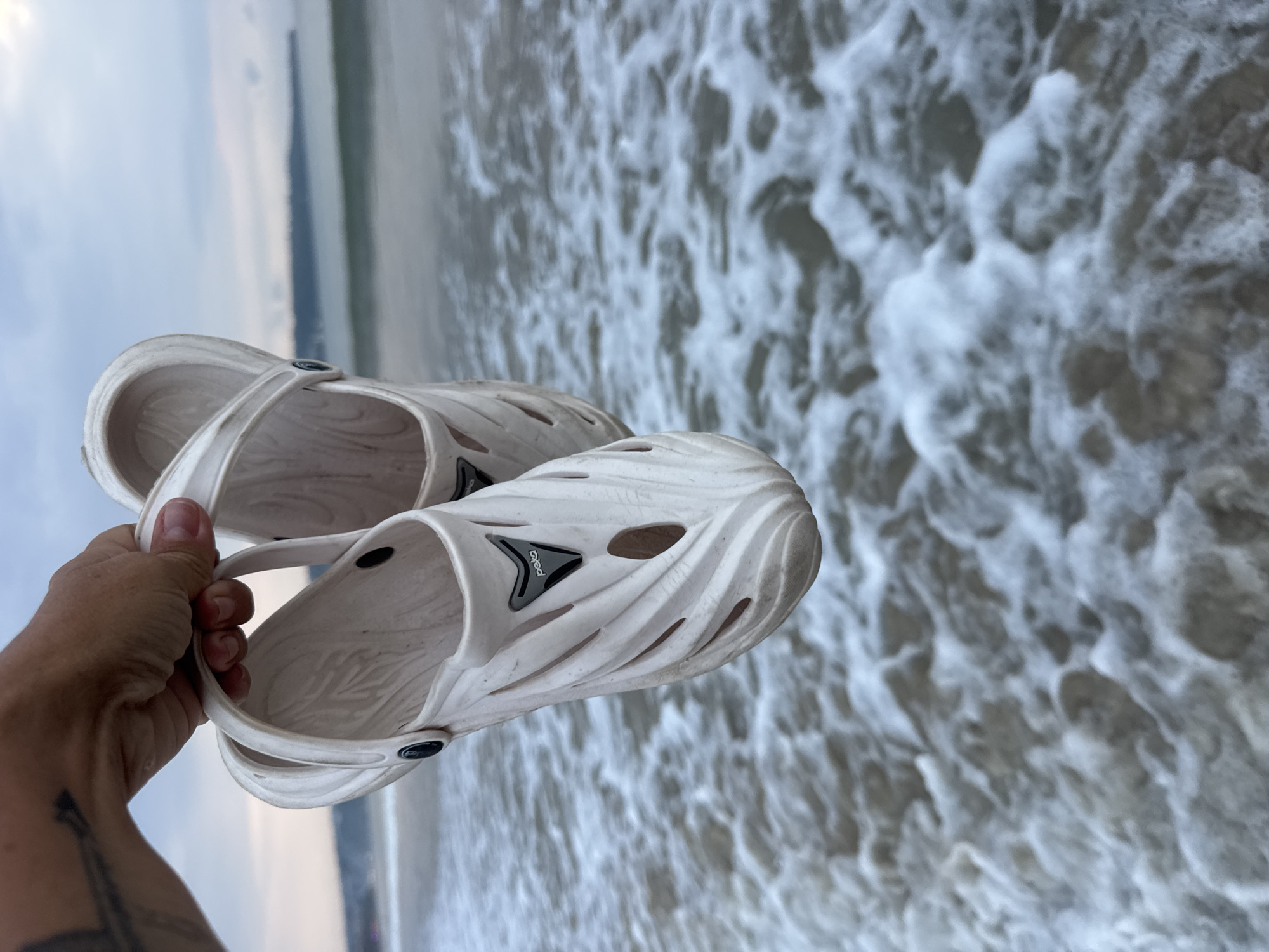 A human hand holds a pair of white, molded plastic clogs above the foamy shallows of Jimbaran Beach, Jimbaran, Indonesia. The hand, visible with tattooed markings on the forearm, clutches the shoes by their attached straps. The clogs feature textured surfaces, multiple perforations, and a black triangular "peka" emblem on the top of the left shoe. The immediate foreground shows churning seawater, receding into a calmer ocean under a pale, overcast sky, with a hazy landmass discernible on the distant horizon.