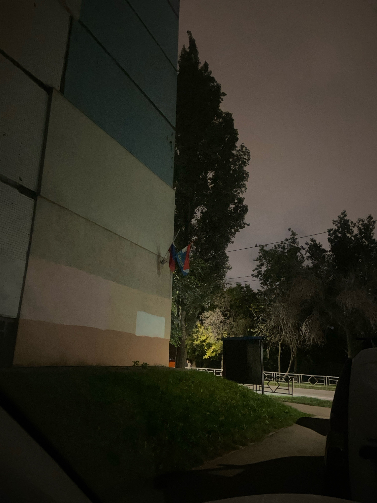 The image depicts an exterior view of a multi-story building at night in Samara, Russia. The building's facade is divided into horizontal sections of various muted colors, including a light blue, a beige, and a light pinkish-brown. A Russian flag is attached to the building's exterior, partially obscured by a tree. The scene is illuminated by ambient light, suggesting it is nighttime. In the background, trees and a dimly lit street with a fence are visible. A parked vehicle is partially visible in the foreground on the right side. The overall atmosphere is dark and quiet.