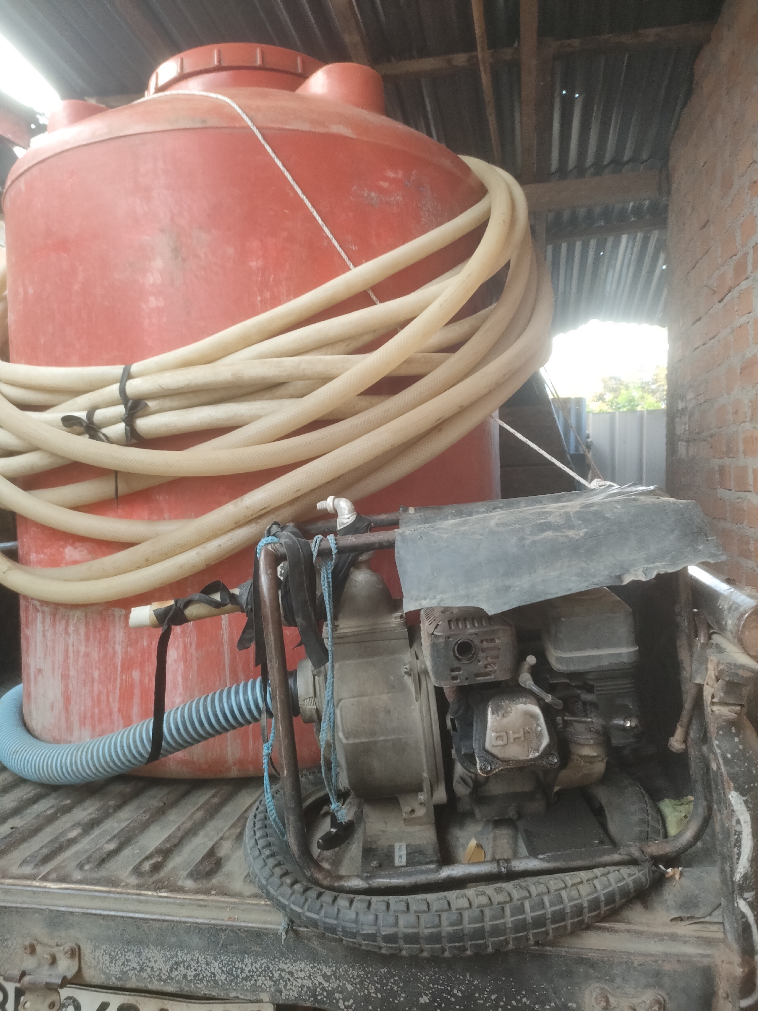 In the city of Padang Dalom, Indonesia, a close-up, eye-level shot captures a red plastic water tank secured on the bed of a vehicle, likely a truck. The tank is large and cylindrical, with a slightly weathered appearance and a screw-on lid. A coiled, off-white hose is draped over the front of the tank, fastened in several places with black ties.
In front of the water tank, a pump is visible. It appears to be a gasoline-powered pump, with a black engine cover and a gray housing. The pump is mounted on a black metal frame, and a spare tire is positioned underneath it, partially obscuring the frame. A blue flexible hose connects from the bottom of the water tank to the pump. Several black straps and a blue rope are used to secure the pump and hoses.
The background shows a corrugated metal roof supported by wooden beams, suggesting an outdoor or semi-covered area. To the right, a brick wall made of reddish-brown bricks is visible. The overall scene is dusty and appears to be an operational setup for water transport or distribution. The ground beneath the vehicle is not clearly visible, but the texture of the truck bed suggests it is a ribbed metal surface.