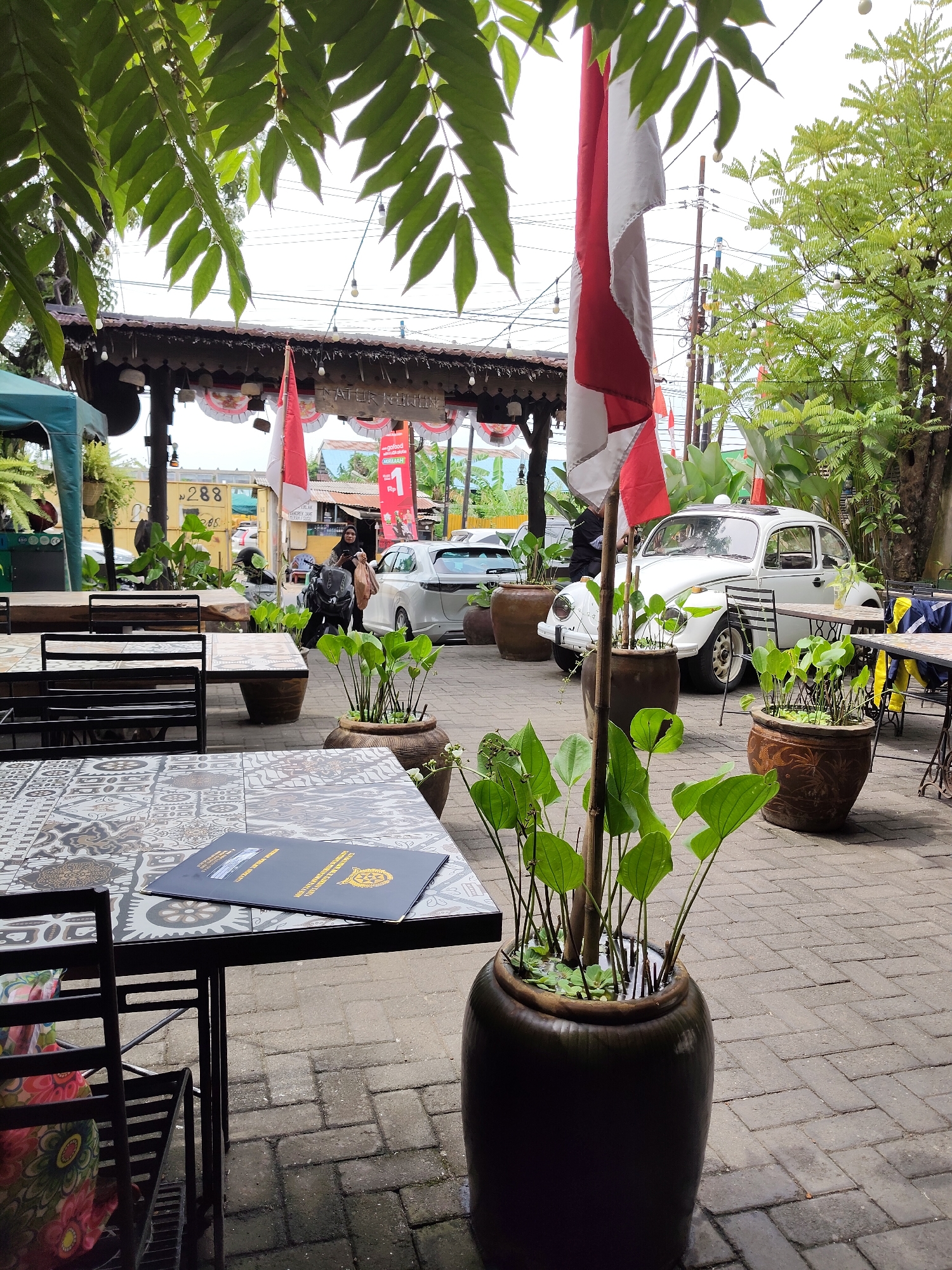 The image depicts an outdoor seating area with tables and chairs on a brick-paved patio. Several potted plants are strategically placed around the area, including one large urn filled with water and aquatic plants in the foreground. A red and white Indonesian flag is prominently displayed, hanging from a flagpole. In the background, a white Volkswagen Beetle is parked, and a woman on a scooter is visible near a building with a traditional thatched roof. Various signs and banners suggest a commercial establishment, possibly a restaurant or cafe, is located here. The overall atmosphere is casual and relaxed, with lush greenery contributing to a pleasant ambiance. This scene is situated in the City of Medan, Indonesia.