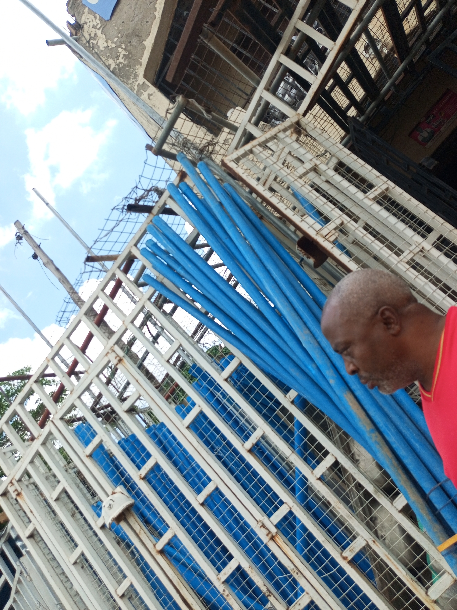 The image shows a man in a red shirt standing in front of a white metal structure with blue pipes behind it. The man is looking down and to his right. The metal structure appears to be part of a construction or storage area, with a checkered pattern and some rust. The blue pipes are cylindrical and are stacked vertically. In the background, there is a cloudy sky and part of a building. The image is taken from a low angle, looking upwards. The scene is likely outdoors, possibly in an industrial or construction site in Arawa, Nigeria.