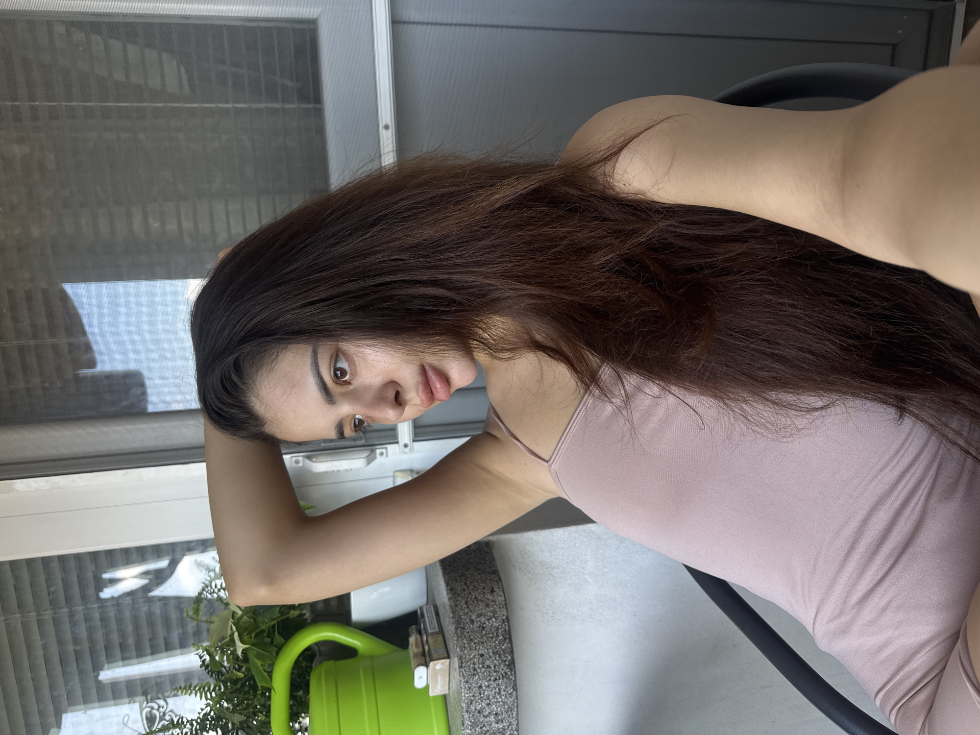The image, originally rotated clockwise, shows a young woman looking directly at the camera. She has long, wavy dark brown hair that cascades over her left shoulder and chest. Her skin is fair, and she has dark eyebrows and brown eyes, with a neutral, slightly contemplative expression. She is wearing a light pink or mauve thin-strapped camisole. Her left arm is bent, with her hand resting on her head, while her right arm is also bent, likely holding the device for the selfie. She appears to be seated on a black chair, partially visible behind her back and arm.
The setting is an indoor space, likely a balcony or a room with a large glass sliding door or window. Behind her, the glass door features prominent vertical and horizontal bars along with a mesh screen, reflecting bright daylight and outdoor scenery. To her left, at the bottom of the frame, a bright green watering can is visible next to lush green potted plants. To her right, on a granular-textured counter or ledge, several small boxes are placed, with the text "Djarum Black" partially readable on one. The bright lighting suggests it is daytime. No specific location cues for Plovdiv, Bulgaria, are visible in the image itself.
