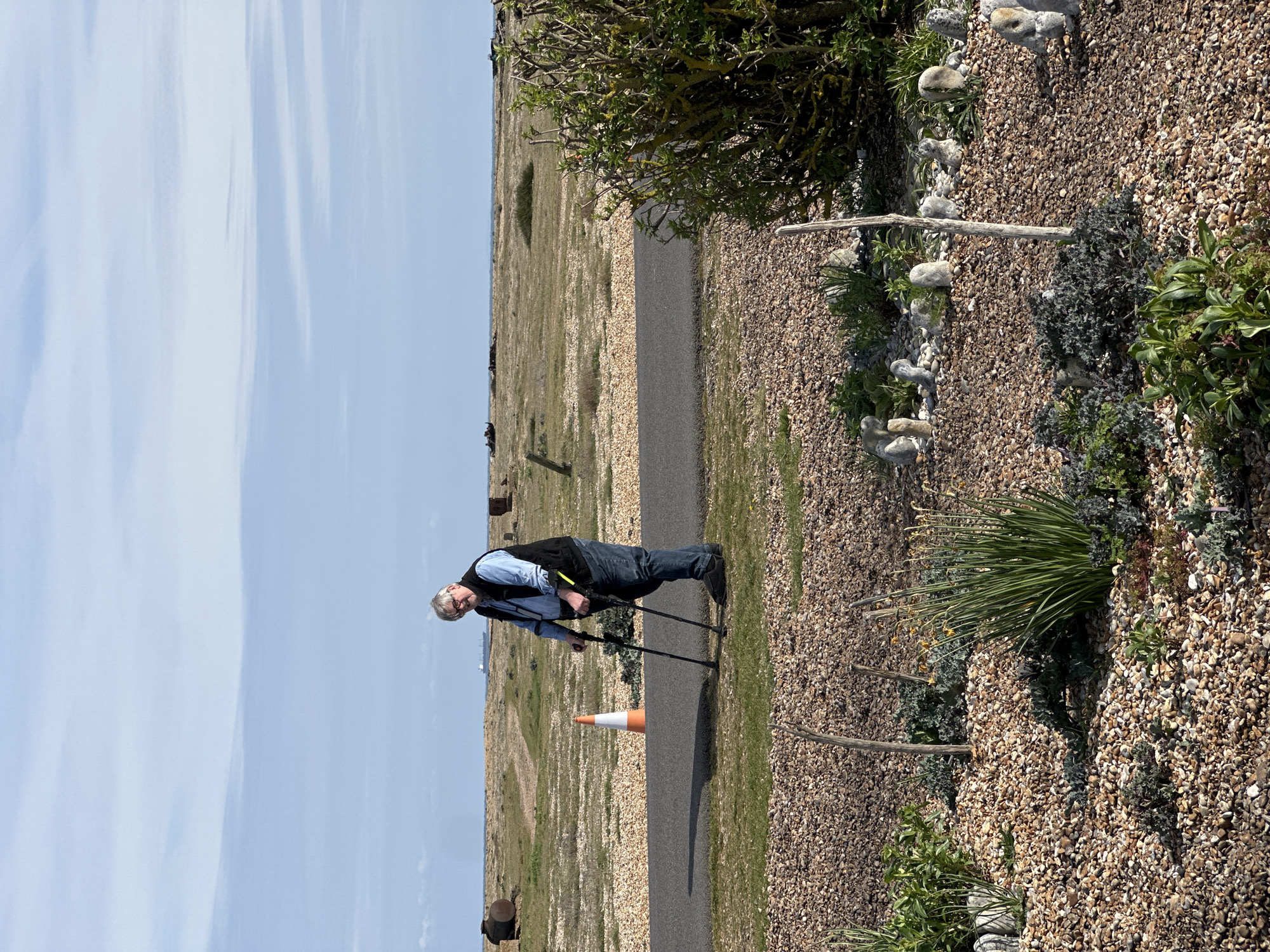 An eye-level shot shows an older man with a cane standing on a gravel path that runs along a landscape. The man has white hair and glasses, and he is wearing a blue shirt, a black vest, and jeans. He is holding two black canes, and he appears to be looking to his left. To the right of the path is a gravel garden with small shrubs and plants. Small painted white stones line the border of the garden. Beyond the path, the landscape stretches out to a distant horizon, with grassy patches among the gravel. A bright blue sky with thin clouds is visible above. An orange traffic cone sits in the path a short distance from the man. The scene suggests an outdoor setting, potentially a garden or park, in the Folkestone and Hythe District of the United Kingdom.