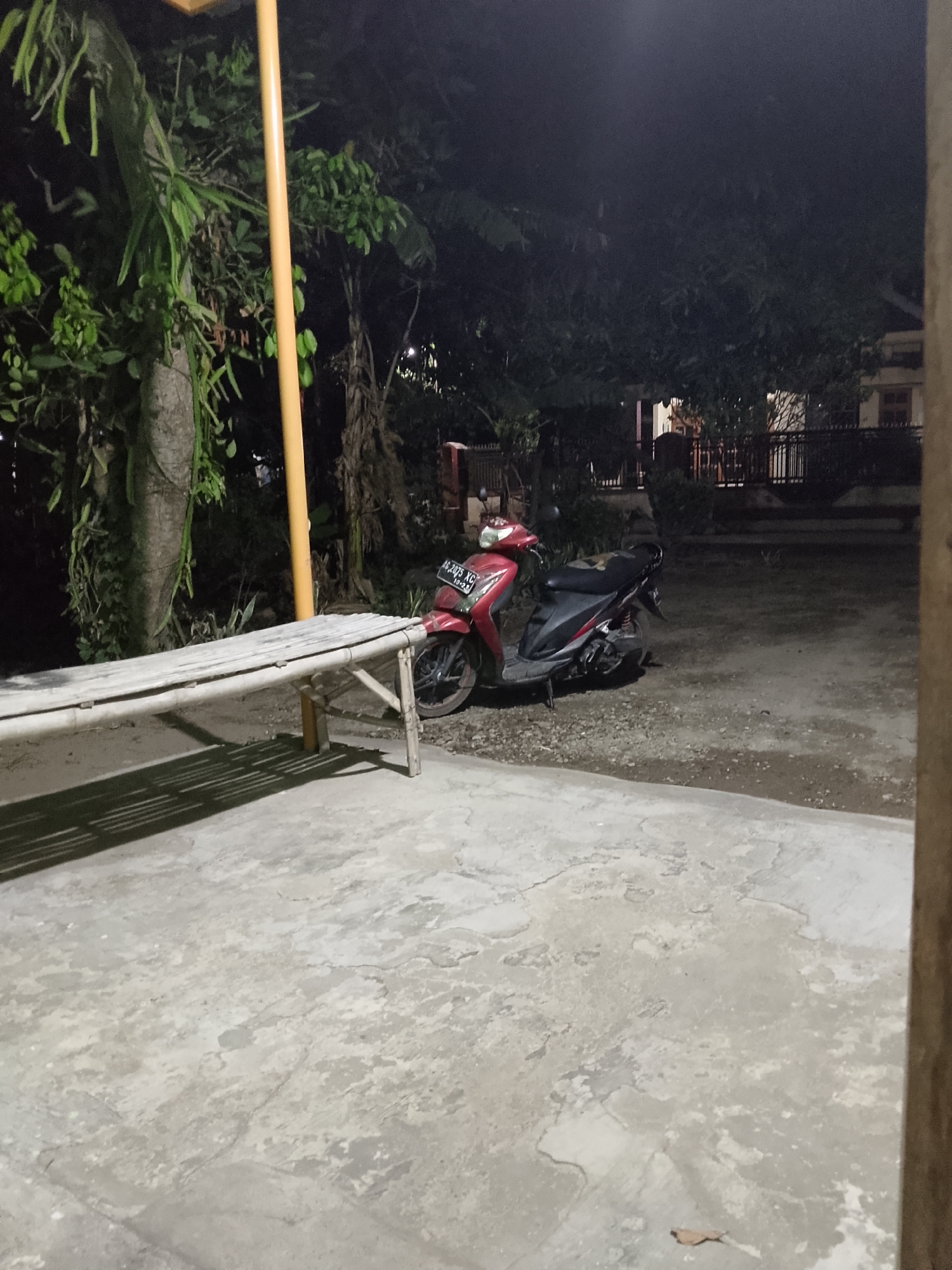 The image captures a nighttime scene in Kelurahan, Indonesia, focusing on an outdoor area with a concrete ground and a dirt and gravel section. A red and black scooter is parked on the dirt area, its license plate clearly visible with the characters "AG. 2075 XG". To the left of the scooter, a long bamboo bench is positioned on the concrete. Behind the bench, a large tree with vines growing on it stands tall, illuminated by an unseen light source, and a yellow pole is visible. Further in the background, a fence with metal railings separates the yard from a building with several windows. The environment is dimly lit, typical of nighttime in a residential area. There are no people present in the frame.