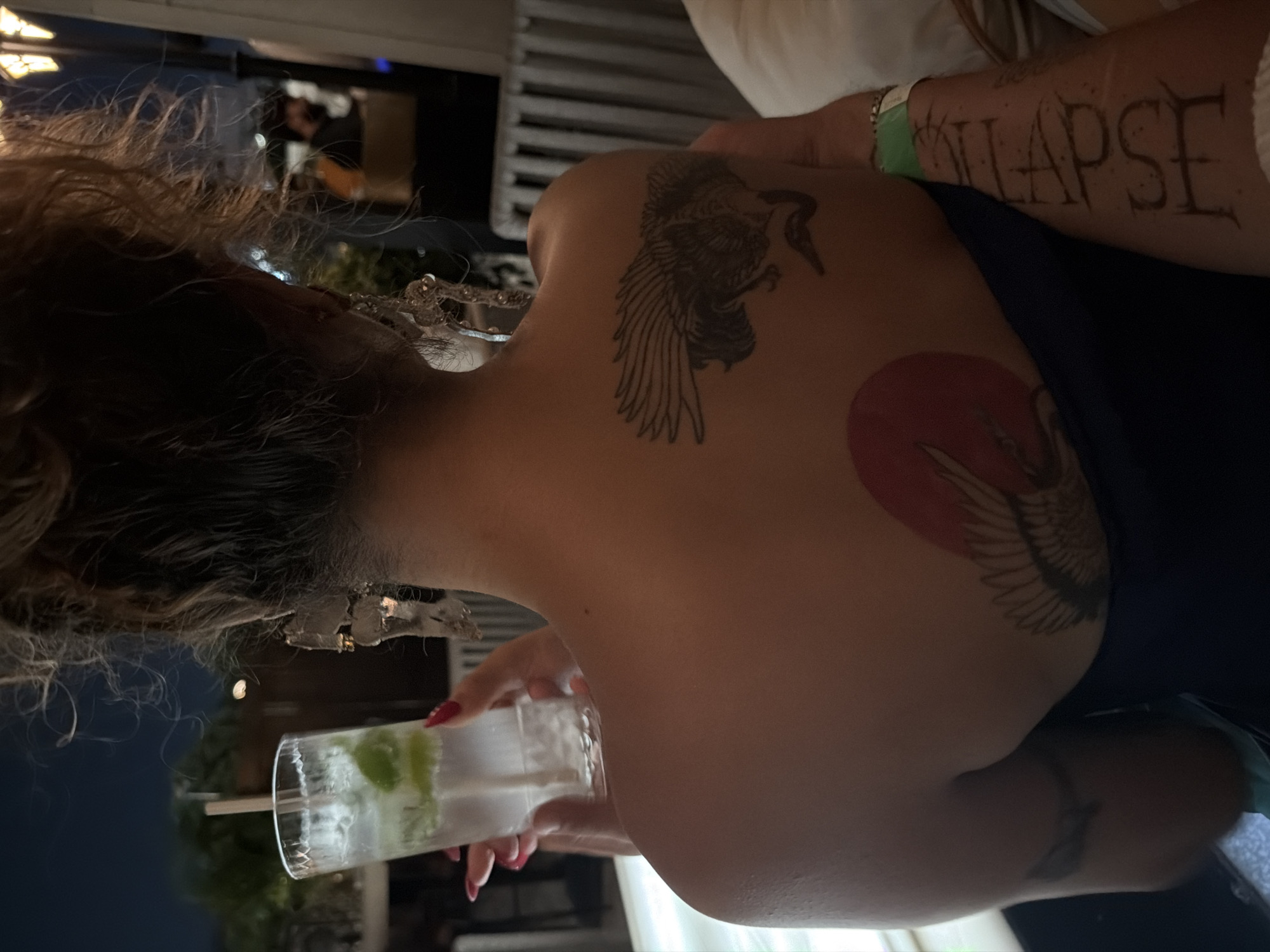 An individual, viewed from behind, is holding a clear glass with a beverage and ice, and what appears to be lime slices. The individual has long, curly, blonde hair. They are wearing large, ornate earrings and a dark blue strapless top. A tattoo of a bird with outstretched wings is visible on the upper back. Below this, there is a tattoo of a bird with a red circle behind it. On the individual's forearm, a tattoo reads "ILAPSE". The scene appears to be outdoors at night, possibly a restaurant or bar setting, with blurred background elements suggesting other patrons and lighting. The location is identified as New Delhi, India.