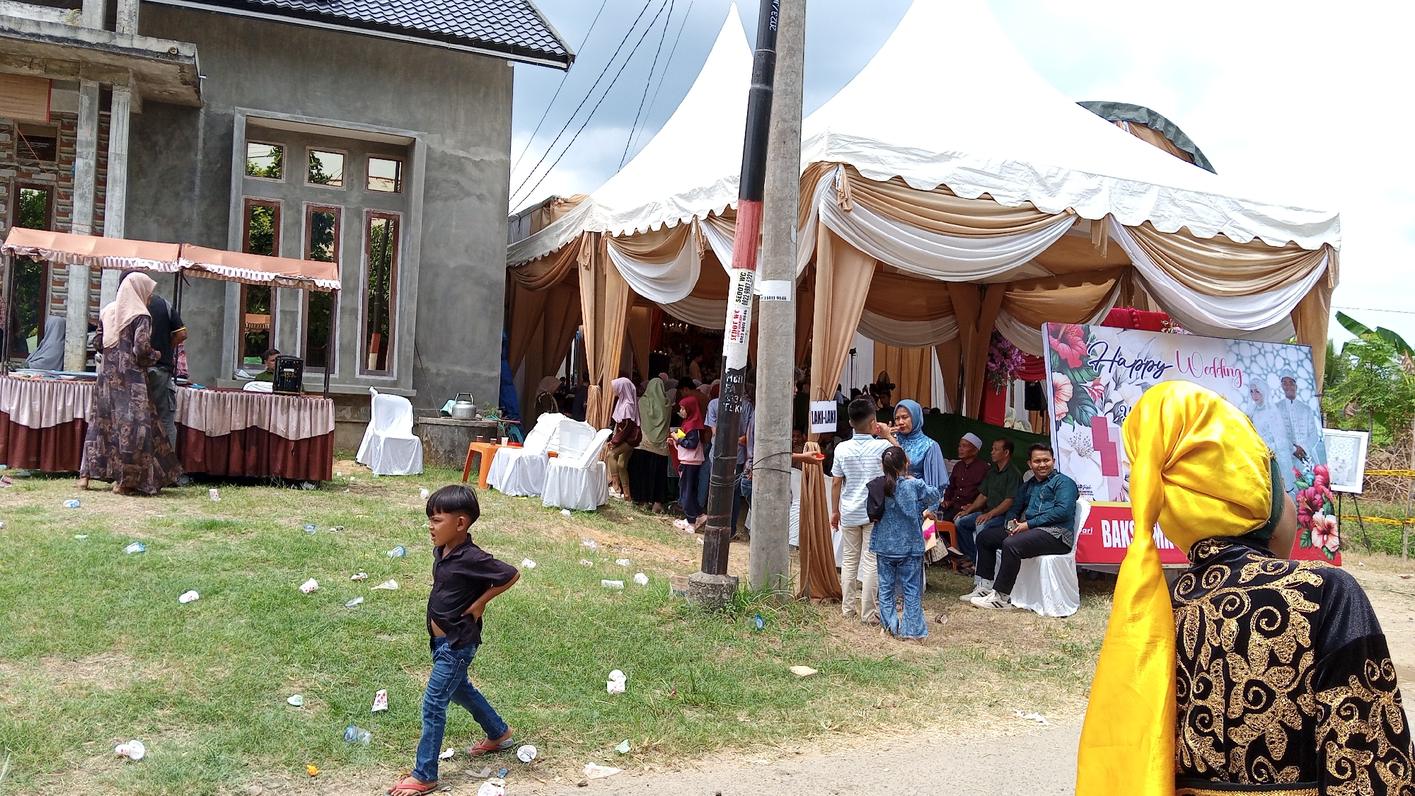 This is an outdoor photograph taken in the city of Payacut, Indonesia, depicting a wedding celebration. A large, cream and gold tent is set up on the right side of the image, draped with fabric. Beneath the tent, a group of people are gathered, some seated and some standing. A large "Happy Wedding" banner featuring a wedding couple is prominently displayed. To the left of the tent, a portion of a building with grey concrete walls and windows is visible. In the foreground, a young boy in a black shirt and blue jeans walks across a grassy area littered with paper cups and plastic bottles. A table draped with a brown and beige cloth is set up near the building. Another group of people, including women in hijabs and men in formal attire, are visible near the table. The overall environment suggests a festive but somewhat informal gathering.