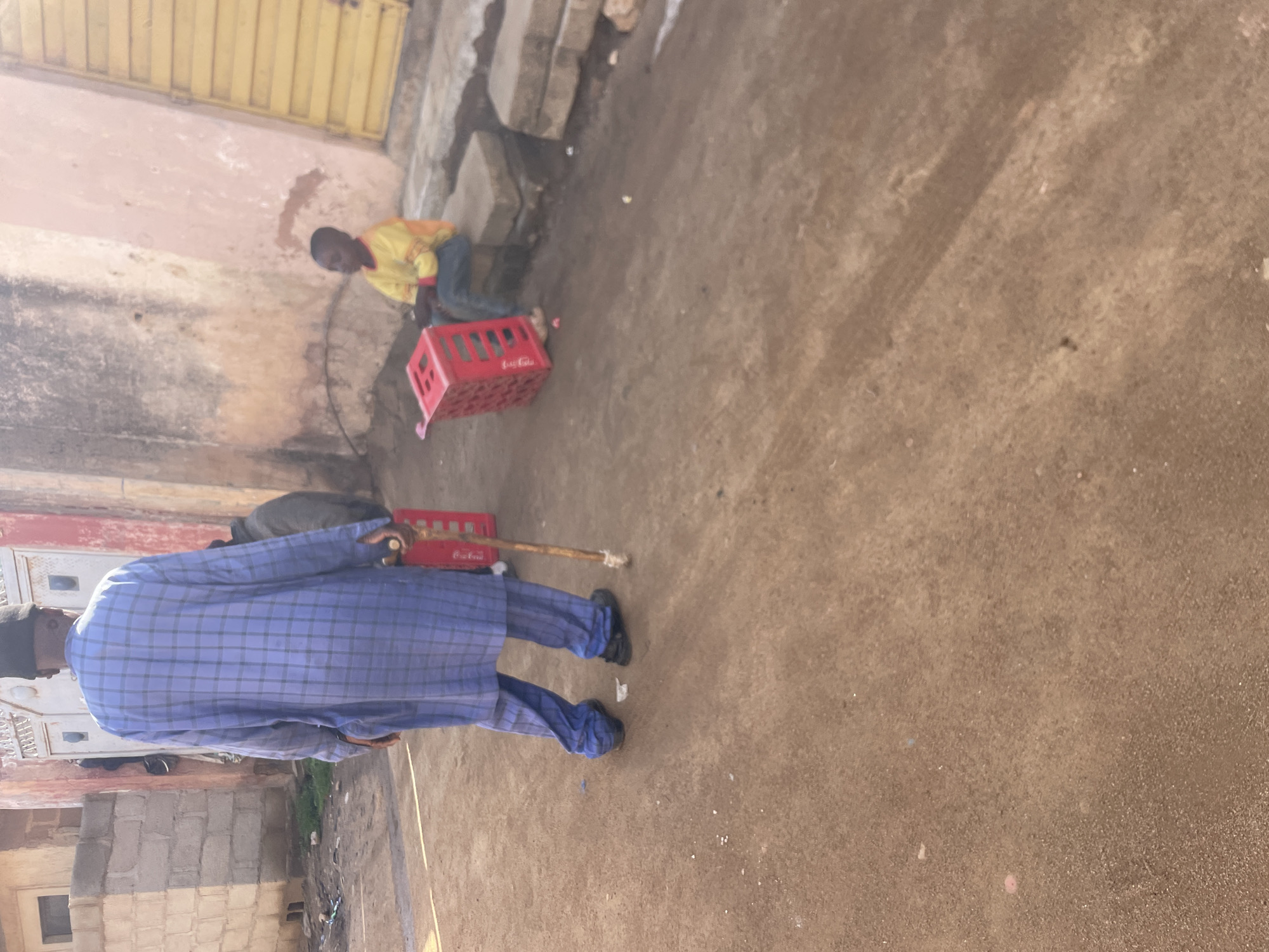In Jos, Nigeria, a man walks with a cane, his back to the camera, wearing a blue plaid robe and dark pants. He is holding a red plastic crate. To his right, a young boy in a yellow and orange shirt sits on concrete blocks next to another red plastic crate. The scene appears to be an outdoor street or pathway with dirt ground and buildings in the background, one of which has a pinkish wall and a yellow door.