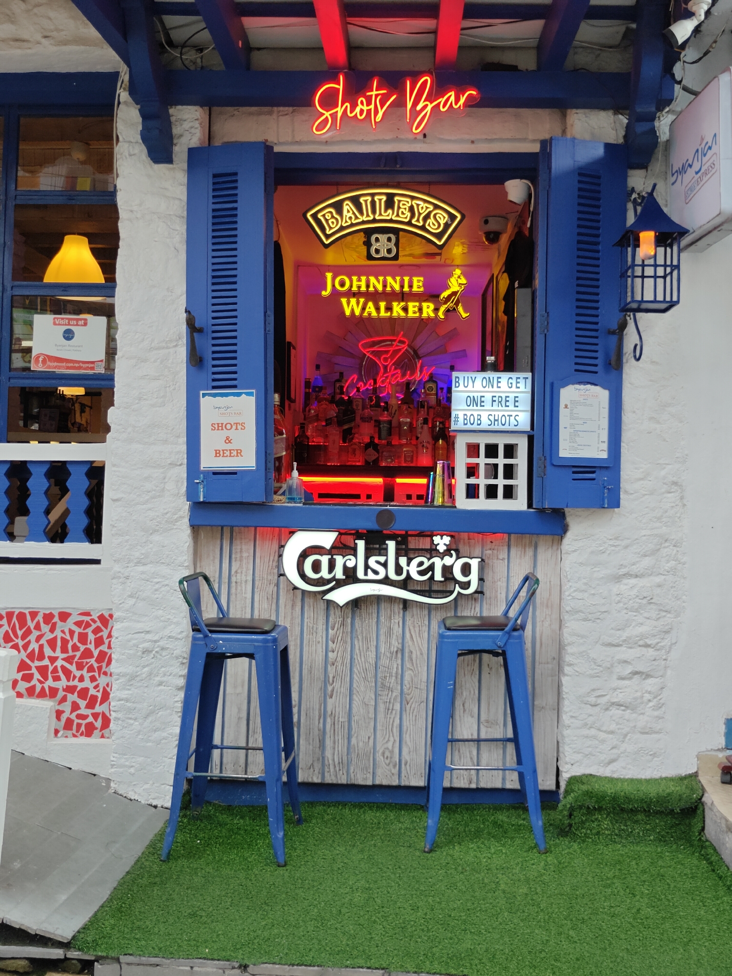 The image shows a close-up of an outdoor bar setup in Pokhara, Nepal. In the center, a window with blue shutters opens to reveal a well-stocked bar area illuminated by neon signs. A neon sign above the window reads "Shots Bar" in red. Inside the bar, illuminated bottles are displayed, with neon signs for "Baileys" and "Johnnie Walker" visible. Below these, a sign advertises "BUY ONE GET ONE FREE #BOB SHOTS". To the left of the bar, a window displays a sign that says "Visit us at Byanjan Restaurant". Two blue bar stools with black seats are positioned in front of the bar counter, which has a "Carlsberg" logo sign on it. The ground in front of the bar is covered in artificial grass. The wall behind the bar is made of white stone, and the building's exterior is painted white with blue accents. A blue lantern hangs to the right of the window. The overall scene suggests a vibrant and inviting establishment.